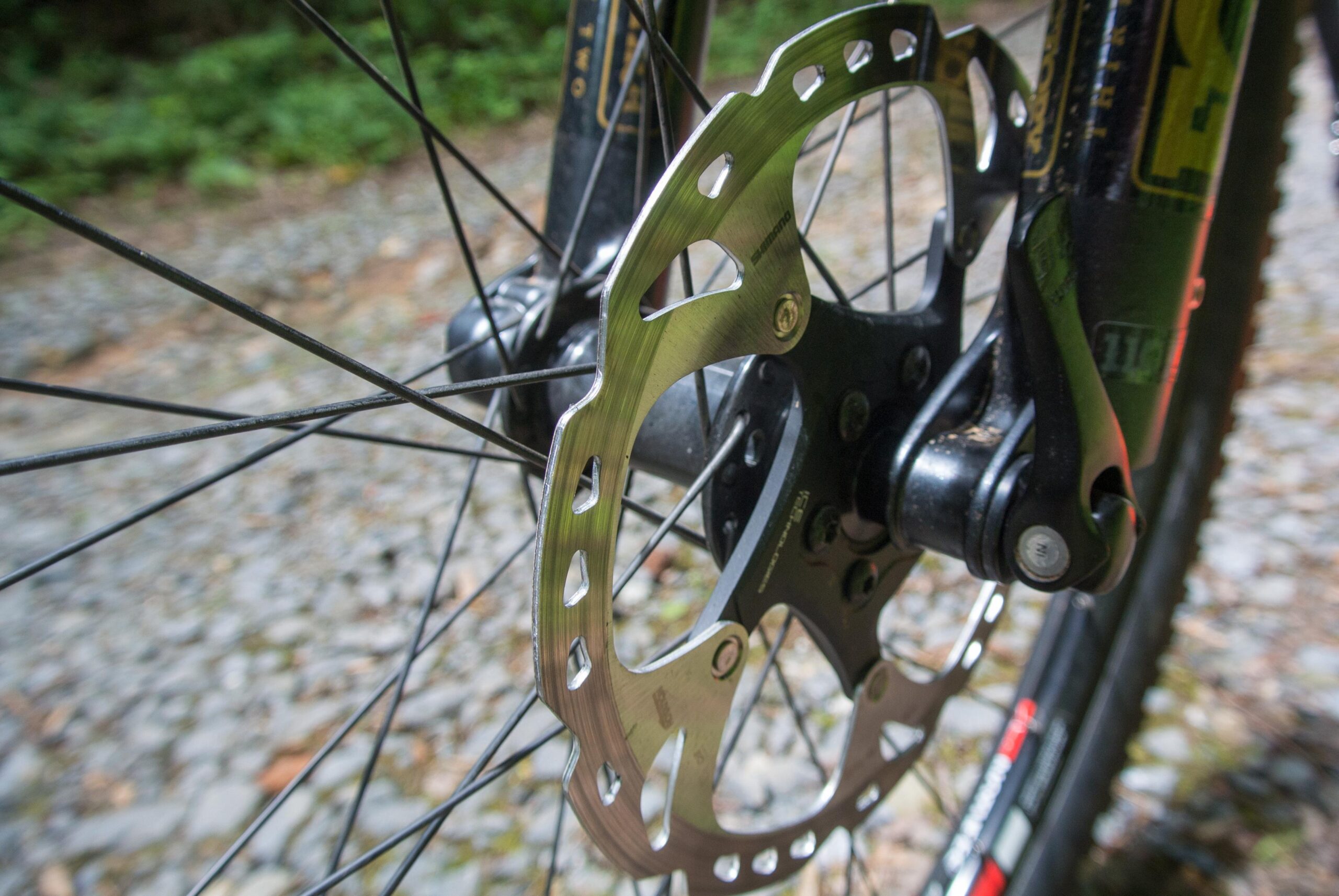 Niner Air 9: Close-up of a bicycle disc brake rotor mounted on a wheel, showcasing the metallic surface and design details. The background features a gravel path and greenery, indicating an outdoor setting.