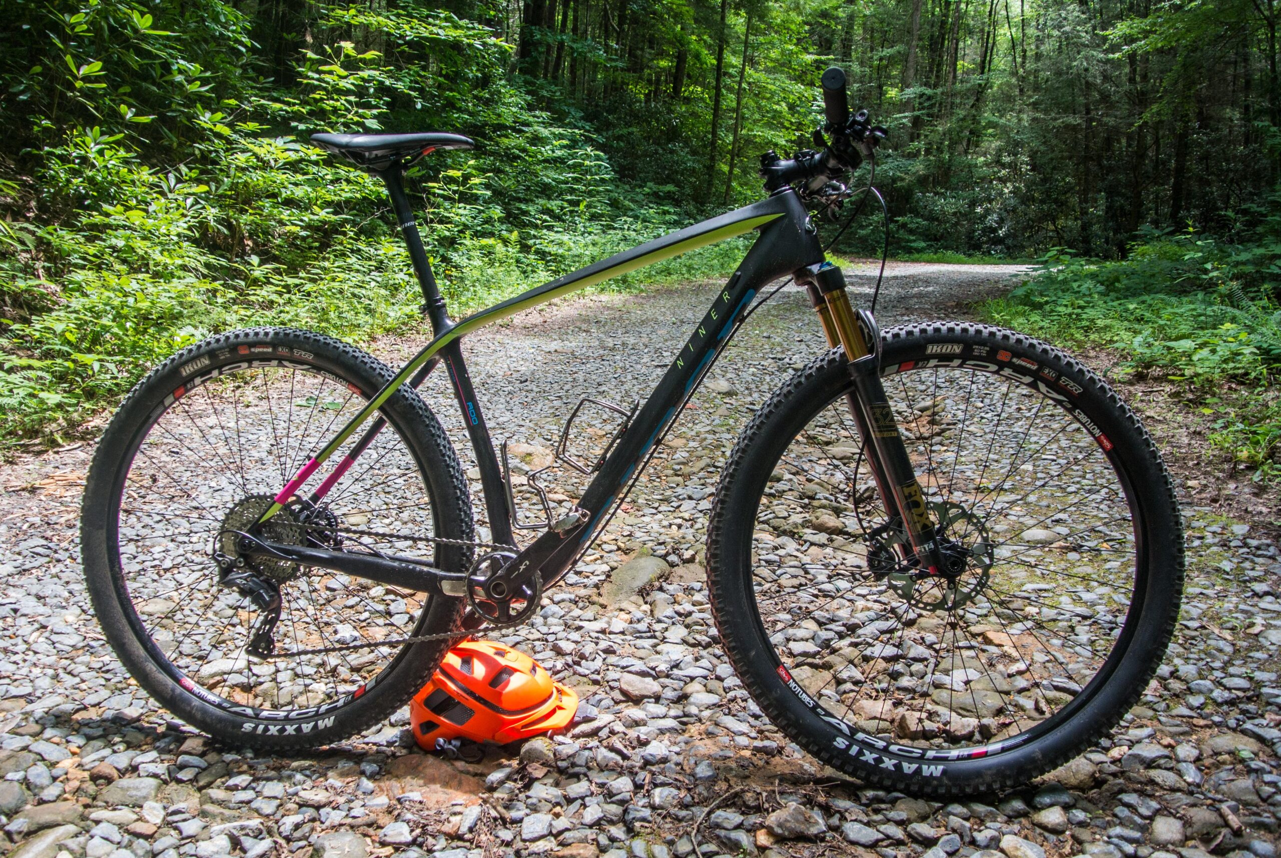 Niner Air 9: A mountain bike with a sleek black and silver frame is parked on a rocky trail surrounded by dense green foliage. An orange helmet rests on the ground beside the bike, highlighting its outdoor cycling context.