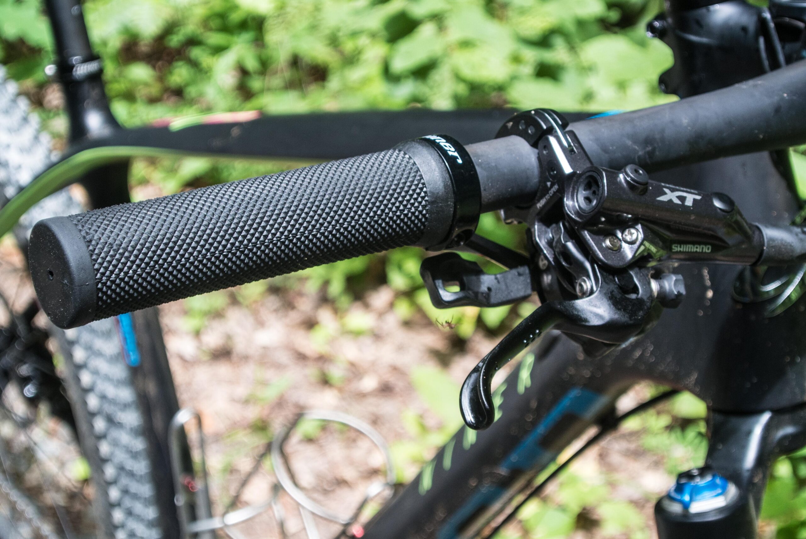 Niner Air 9: A close-up view of a bicycle handlebar featuring a textured black grip, a Shimano XT brake lever, and part of the handlebar itself. The background includes green foliage, suggesting an outdoor setting.