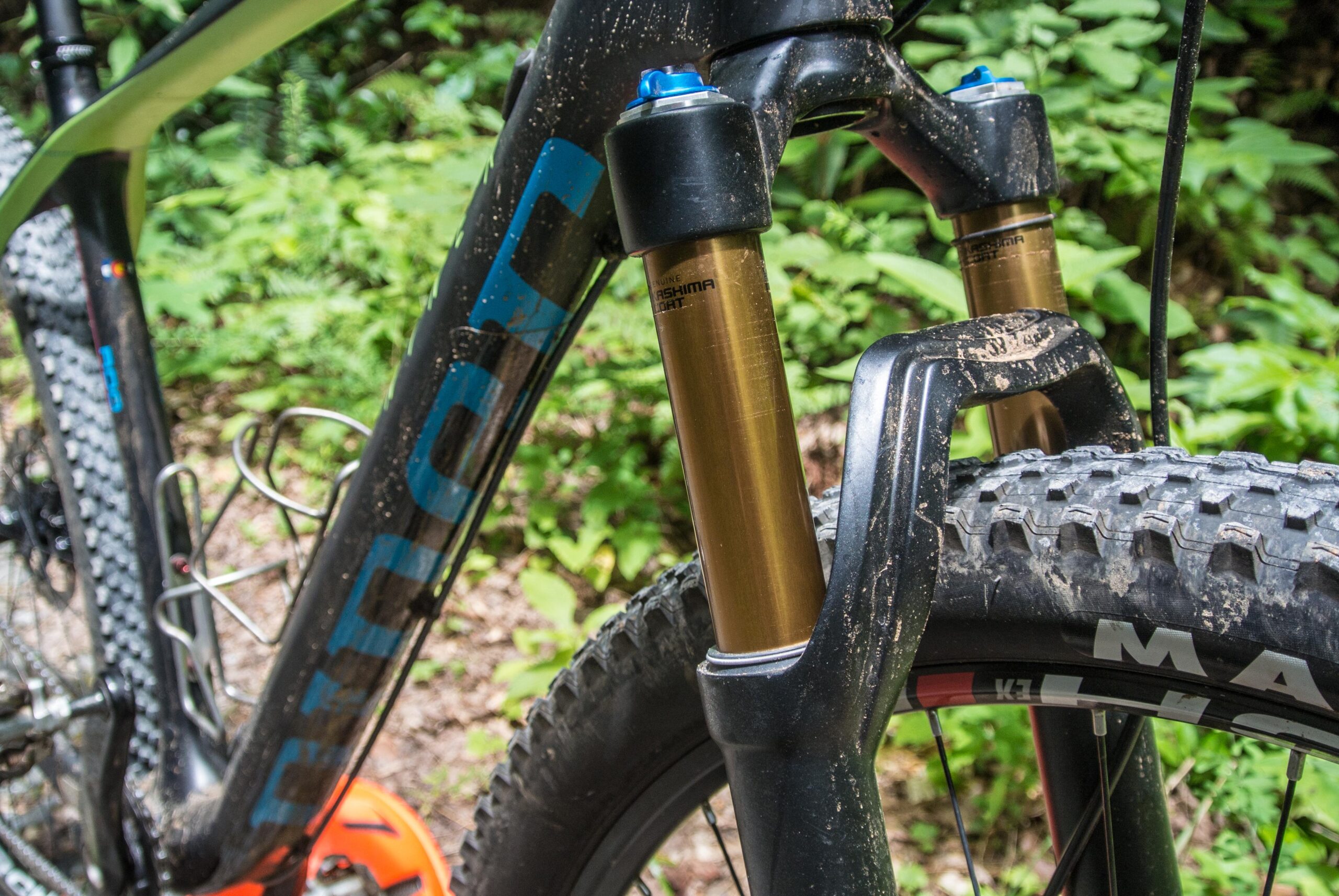 Niner Air 9: Close-up of a mountain bike's front fork and wheel, showcasing the suspension system with gold-colored stanchions and a black frame. The background features lush greenery, indicating an outdoor environment. The bike shows signs of use with some dirt on the frame and tire.