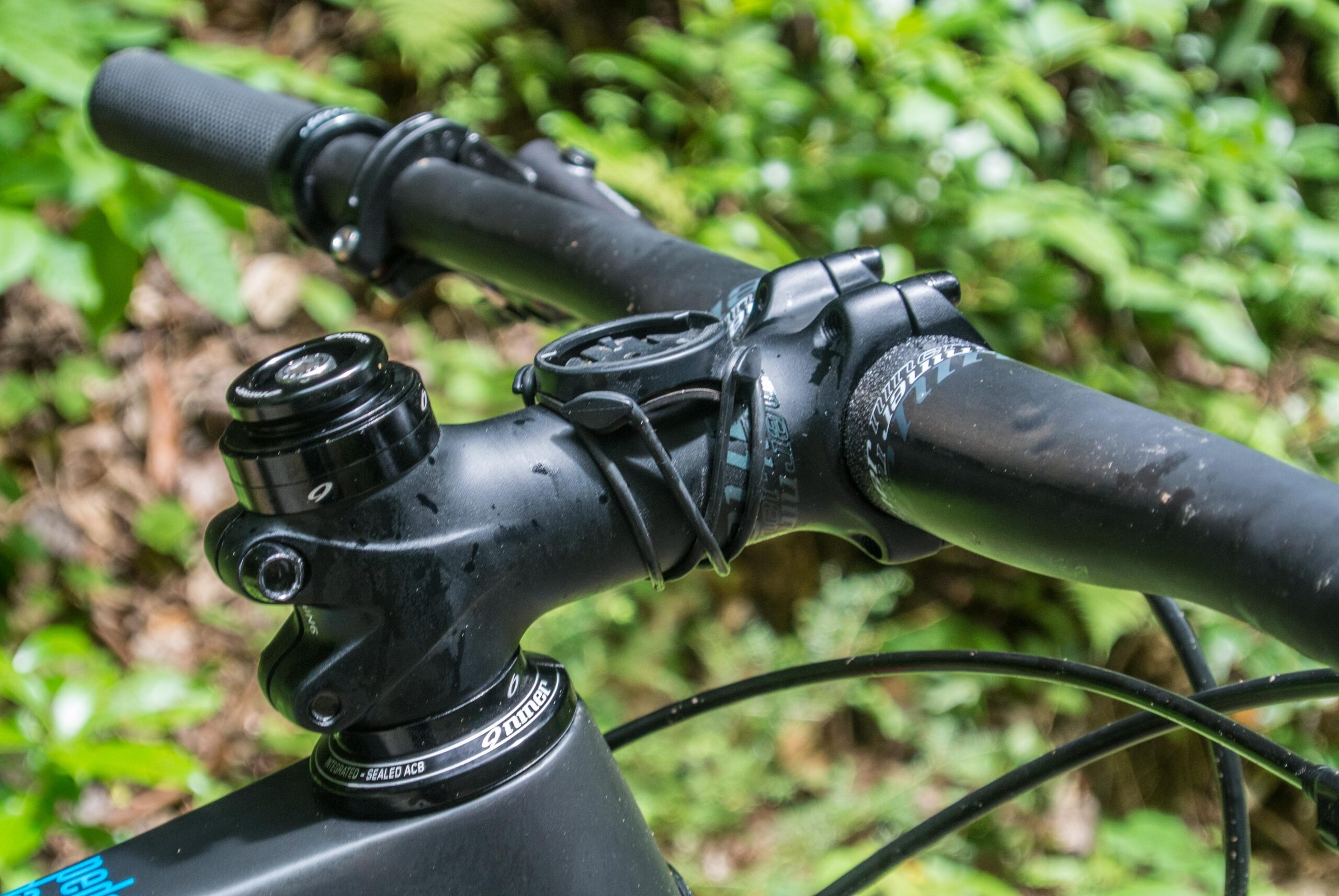 Niner Air 9: Close-up view of a mountain bike handlebar and stem in a forest setting, featuring a black stem, handlebar grips, and a mounted device. The background includes green foliage, creating a natural environment.