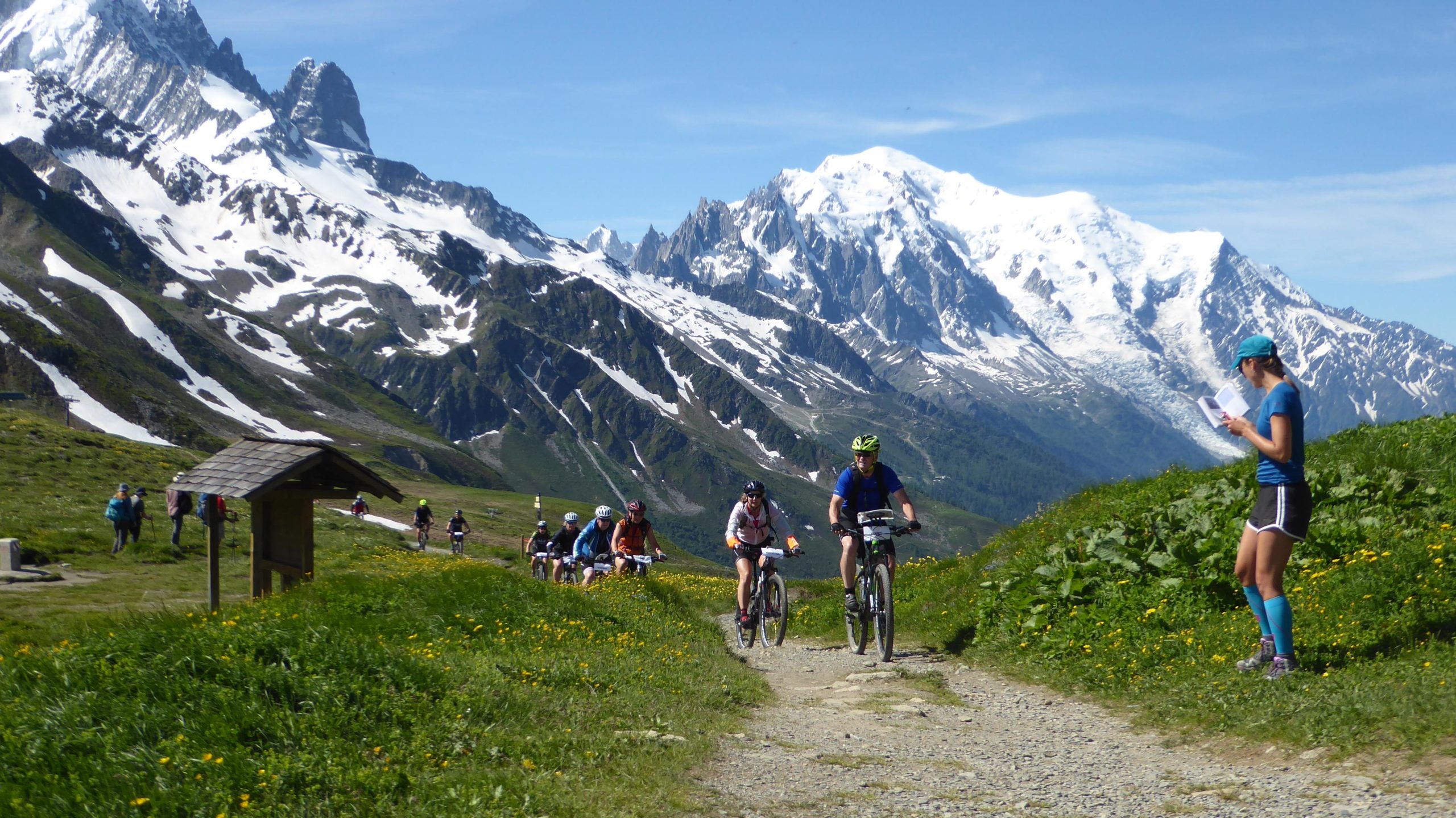 A scenic mountain landscape featuring several cyclists riding on a dirt path, surrounded by lush greenery and wildflowers. Snow-capped peaks rise in the background under a clear blue sky. In the foreground, a woman in athletic attire is reading from a booklet, while more cyclists are seen in the distance. Col de Balme mountain bike trail.