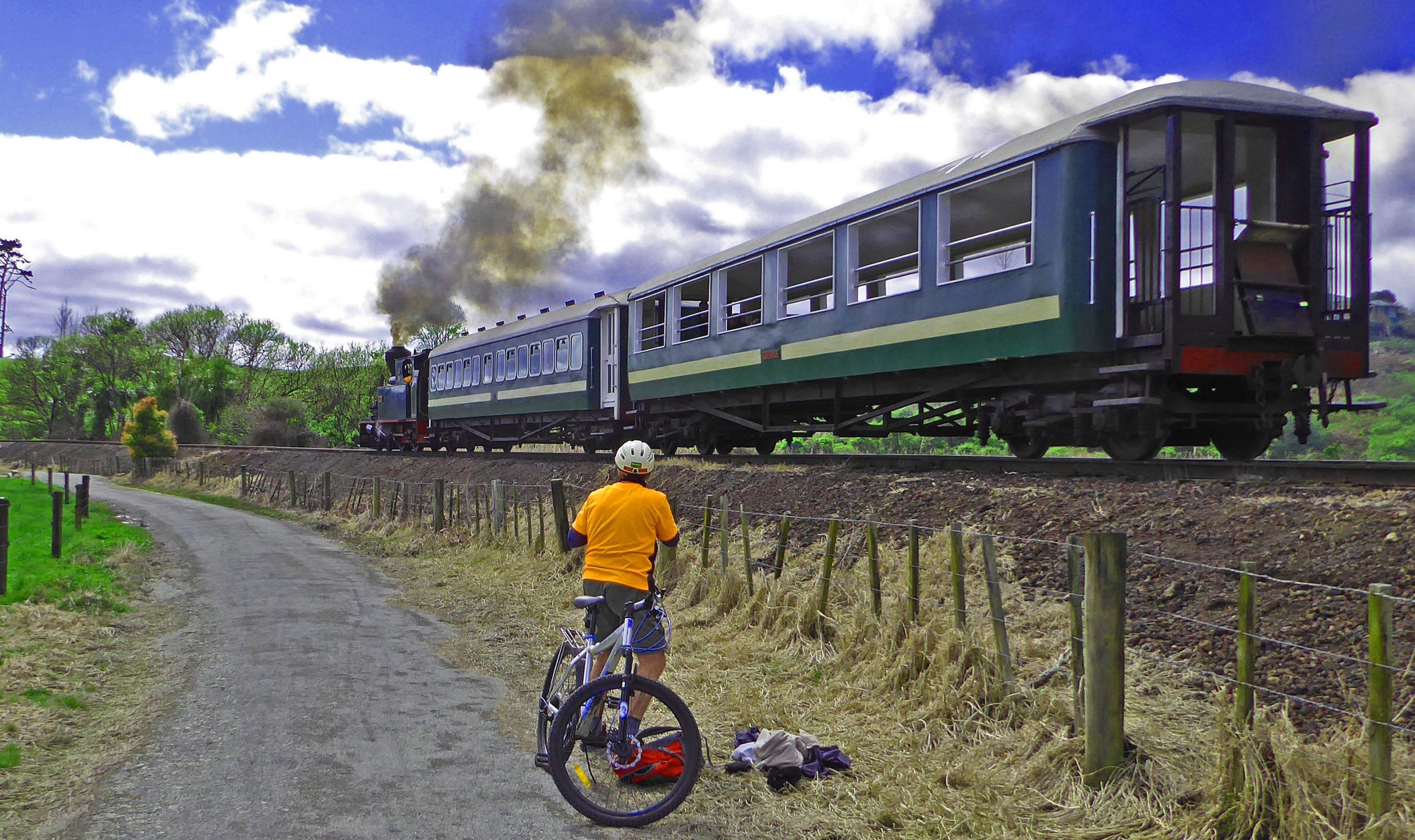 A cyclist in an orange shirt and helmet stands near a dirt path, observing a steam locomotive and passenger train as it travels along the tracks. The scene is set against a backdrop of green trees and a cloudy sky. Smoke billows from the engine, adding to the nostalgic atmosphere. Twin Coast Cycle Trail mountain bike trail.