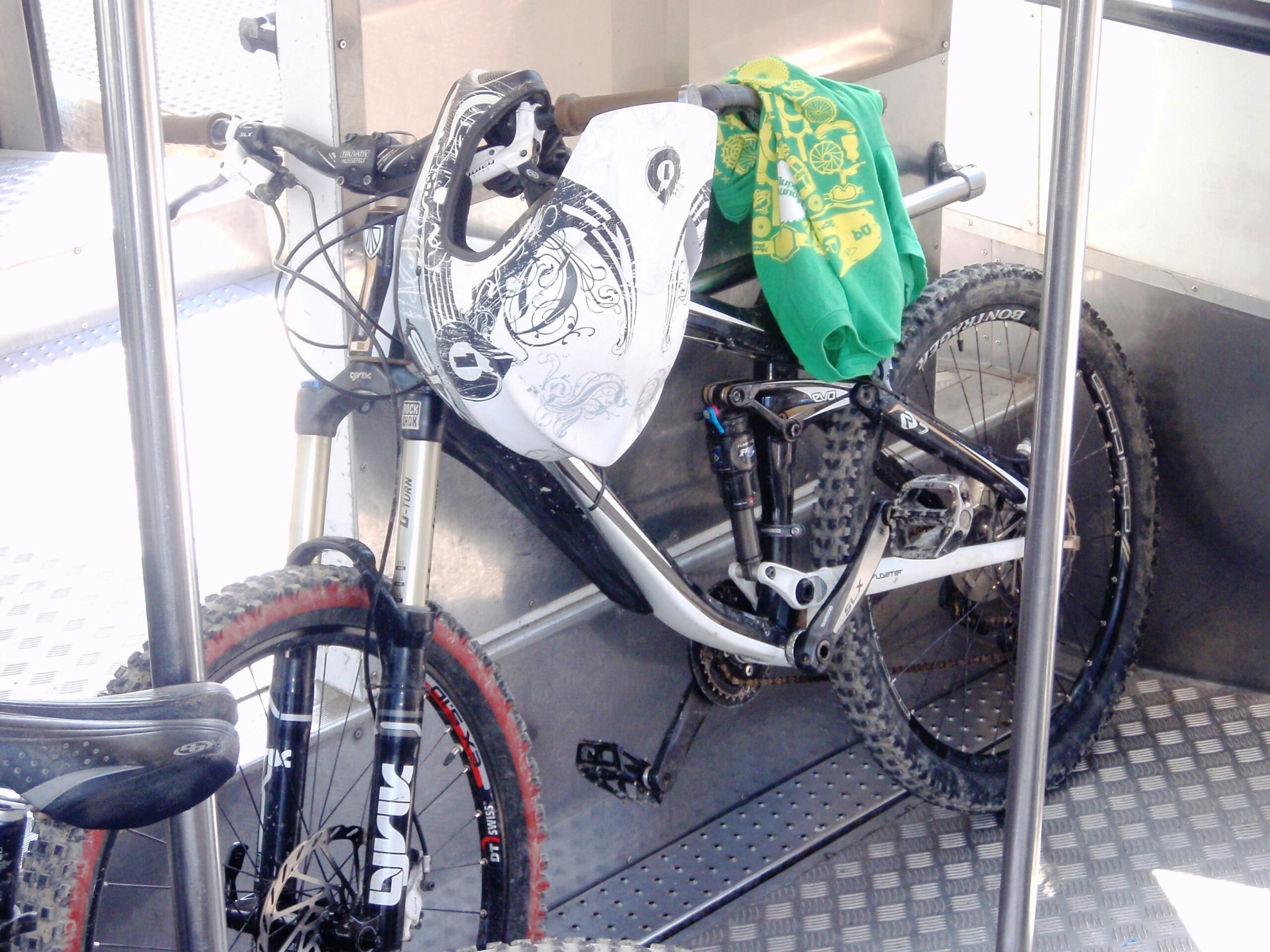 Trek Remedy: A mountain bike with a white helmet rests on a metal floor, with a green shirt draped over the handlebars. The bike has thick tires, a sturdy frame, and visible suspension components. The background shows a sleek interior, likely of a bus or train, with metal poles and a patterned floor.