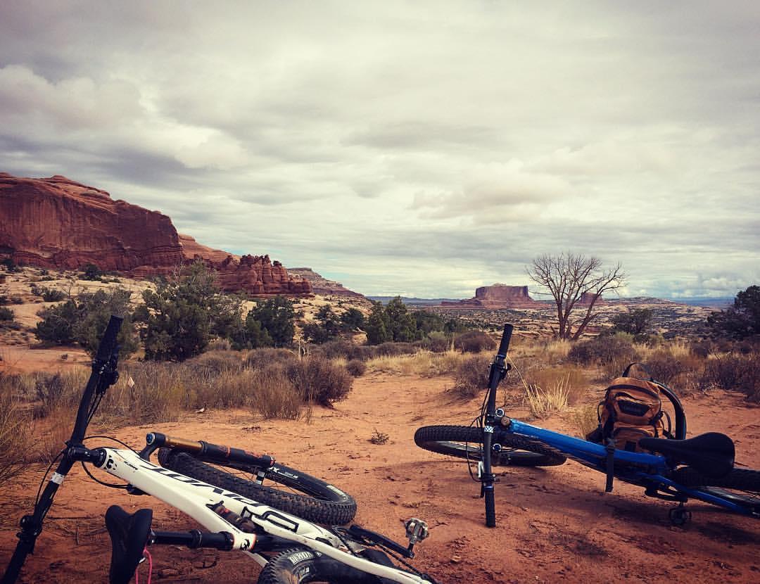 Pivot Trail 429: A scenic view of a desert landscape featuring red rock formations and sparse vegetation. In the foreground, two mountain bikes lie on the sandy ground, with a brown backpack nearby. The sky is overcast with gray clouds, adding a moody atmosphere to the setting.
