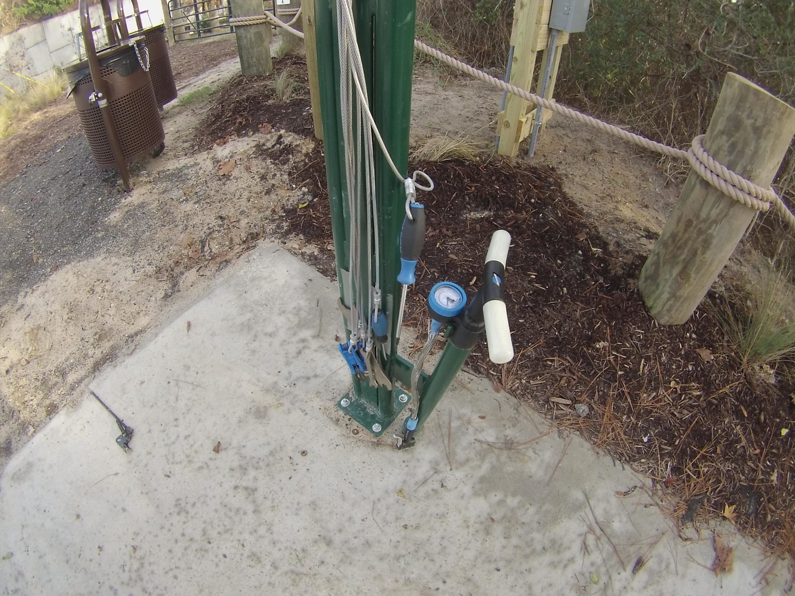 Close-up view of a green vertical pole with various cables and a pressure gauge affixed to it. The pole is set on a concrete slab, surrounded by mulch and grass. In the background, there are two brown trash bins and wooden posts. The image captures equipment possibly related to a recreational facility. Horry County Bike Run Park mountain bike trail.