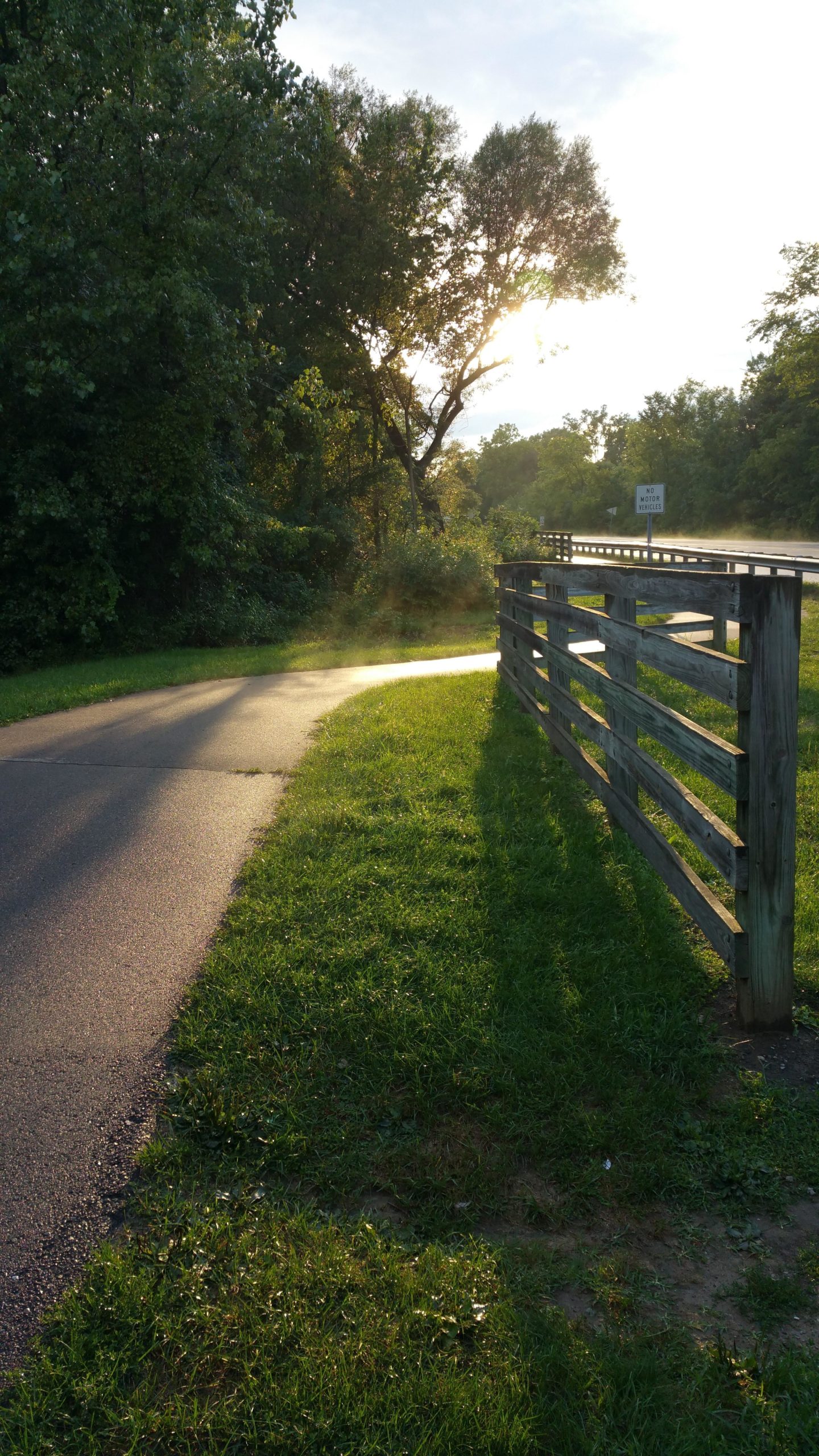 A winding path lined with green grass and trees on one side, leading toward a wooden fence. The sun is setting in the background, casting a warm glow and creating a peaceful atmosphere. A "No Motor Vehicles" sign is visible near the road, indicating that the area is likely intended for pedestrians or cyclists. Hines Park Trail mountain bike trail.