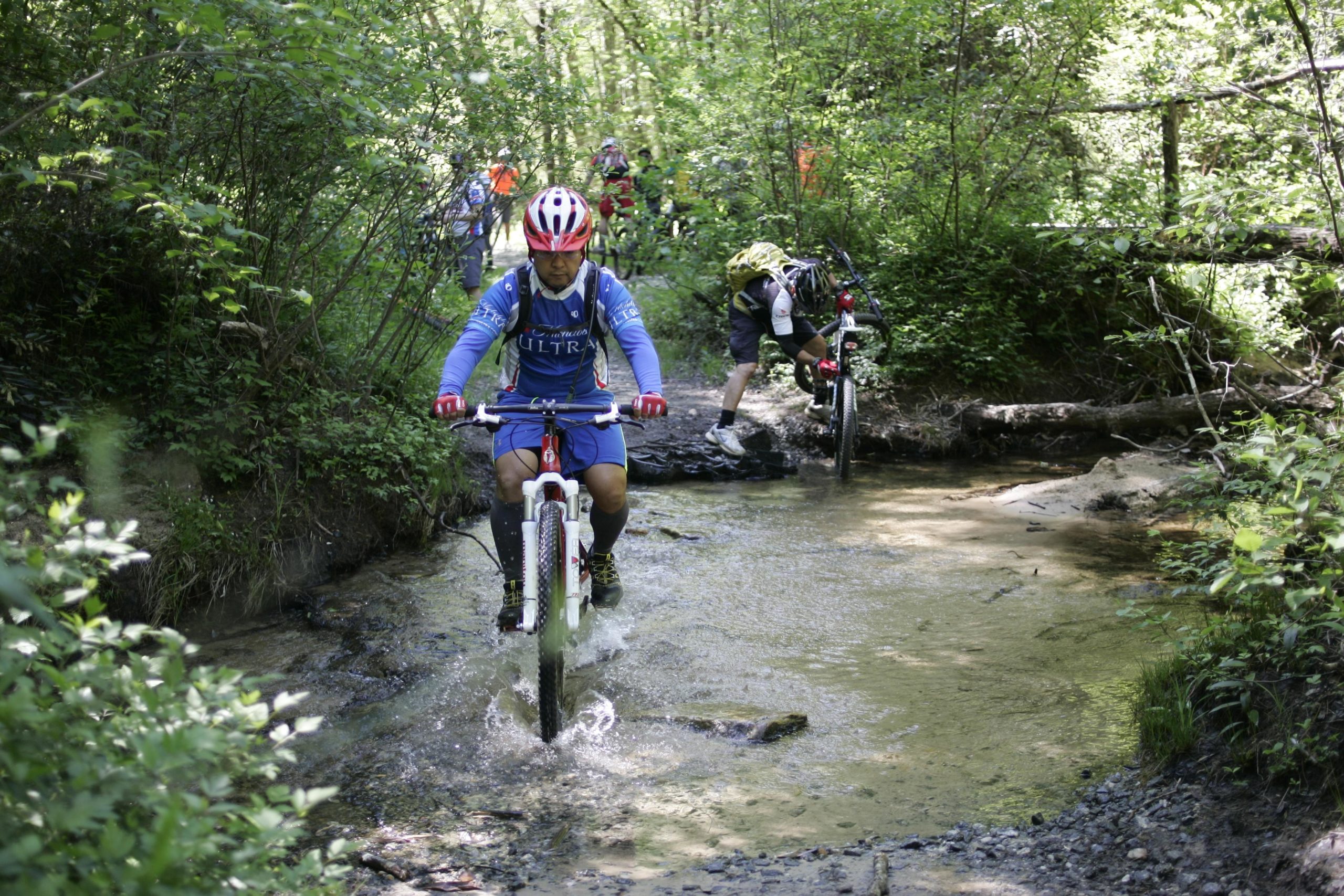 A group of mountain bikers navigating a rocky stream in a lush green forest. One rider in blue is cycling through the water, splashing while others are maneuvering their bikes nearby. Sunlight filters through the trees, creating a vibrant outdoor scene. DuPont State Recreational Forest mountain bike trail.