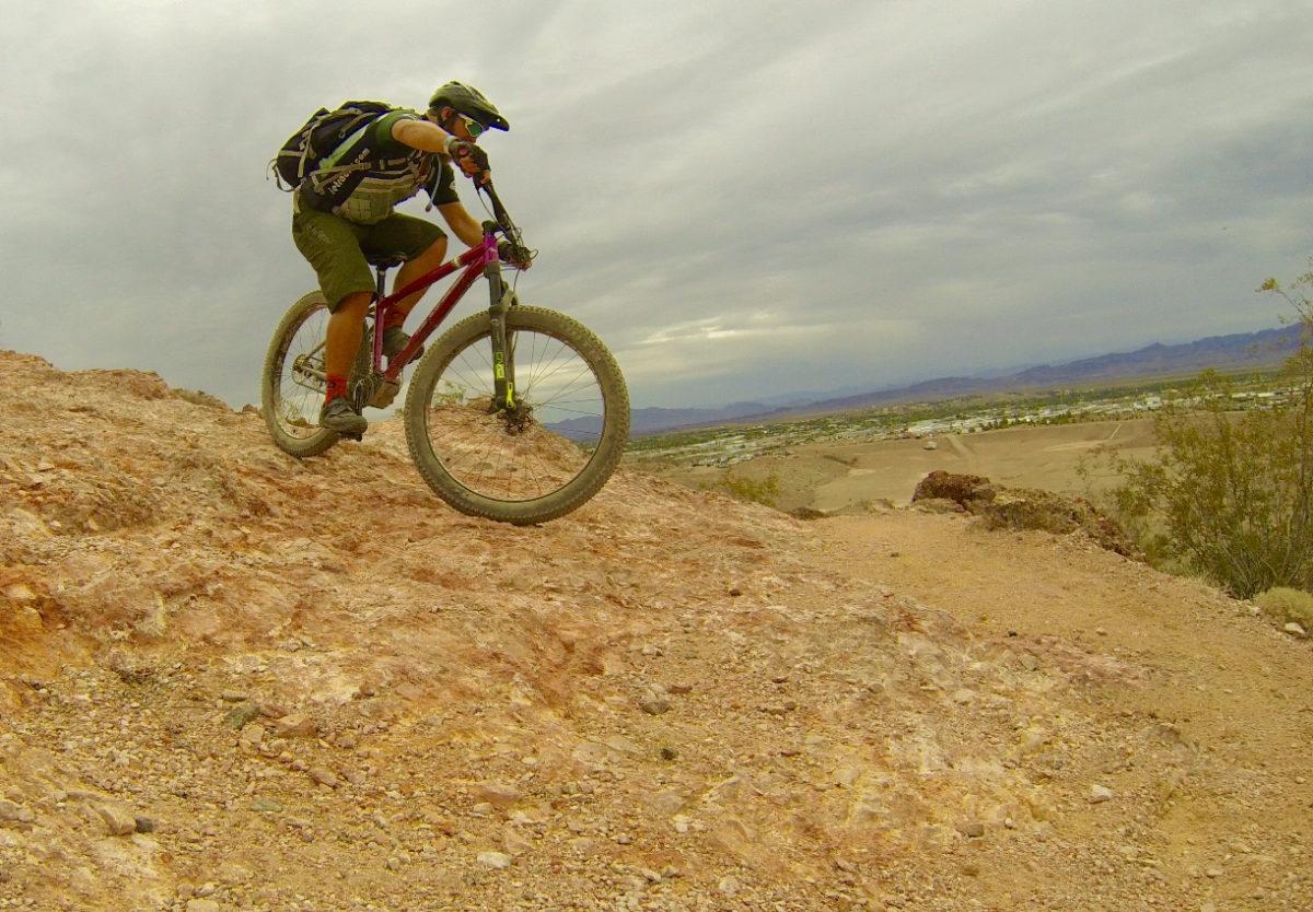 A mountain biker navigating a rocky trail, mid-jump, with a desert landscape and overcast sky in the background. Bootleg Canyon mountain bike trail.