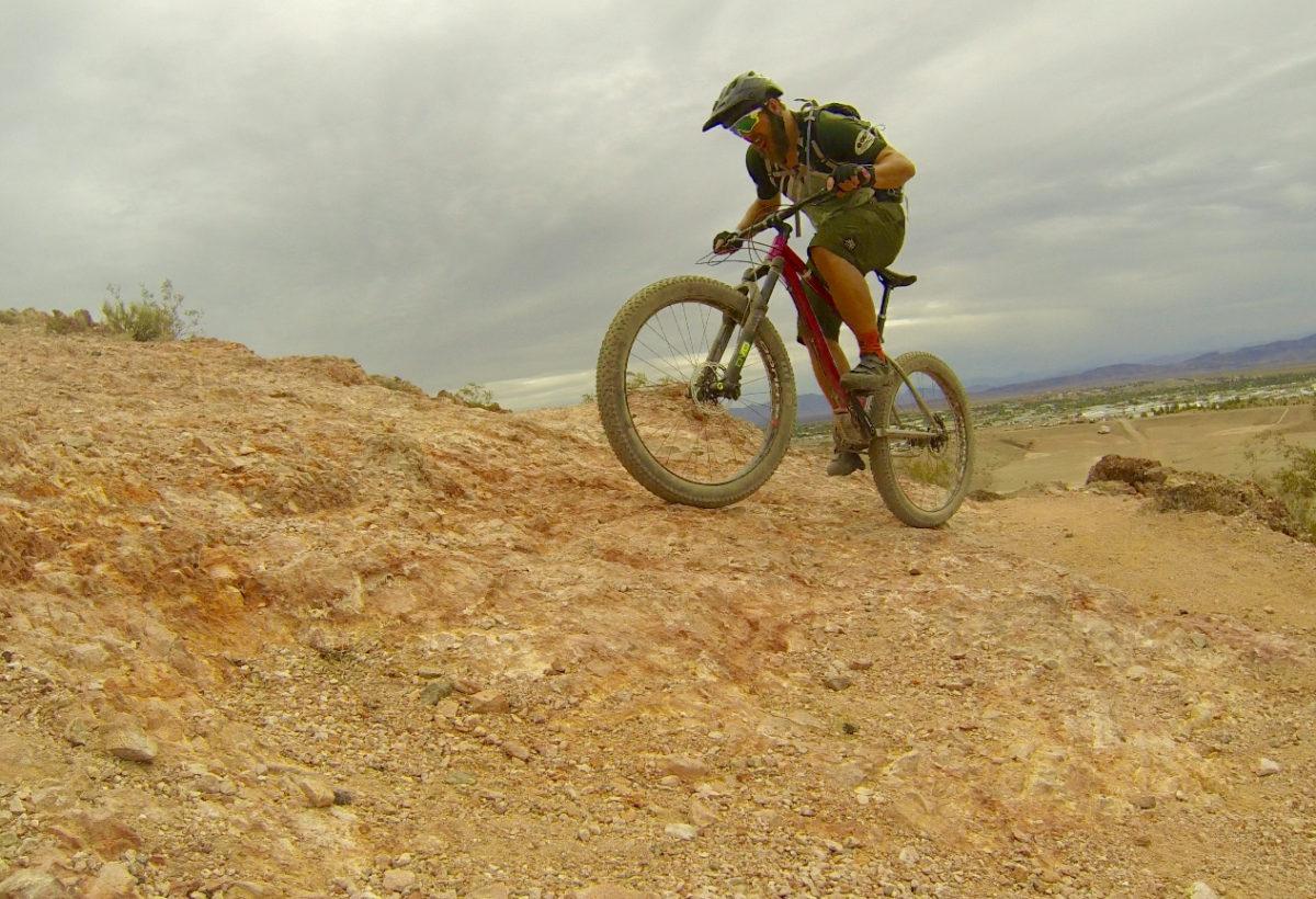 Mountain biker navigating rocky terrain on a bike trail, with a cloudy sky in the background. The rider is wearing a helmet and protective gear. The landscape features hills and sparse vegetation. Bootleg Canyon mountain bike trail.