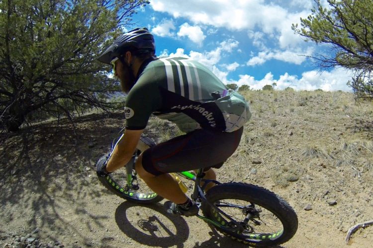 A cyclist riding a mountain bike on a dirt trail surrounded by shrubs and under a partly cloudy sky. The rider wears a helmet and a green and black cycling jersey, focused on navigating the terrain.