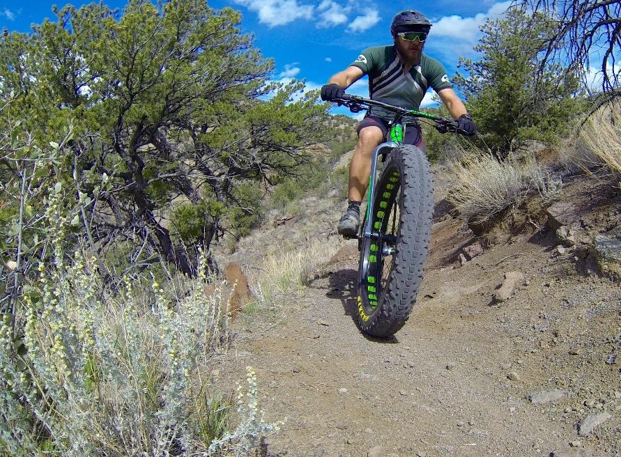 A cyclist riding a fat bike on a narrow dirt trail surrounded by shrubs and trees, with a clear blue sky overhead. The rider is wearing a helmet and sunglasses, showcasing an adventurous stance as they navigate the terrain. Arkansas Hills mountain bike trail.