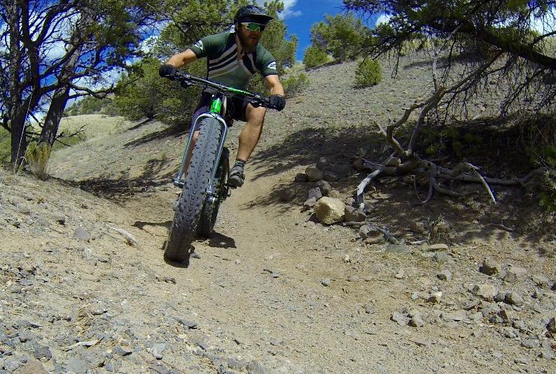 A cyclist riding a fat bike on a dirt trail surrounded by trees, navigating a rocky path under a blue sky with scattered clouds. The rider is wearing a helmet, sunglasses, and a green and black jersey. Arkansas Hills mountain bike trail.