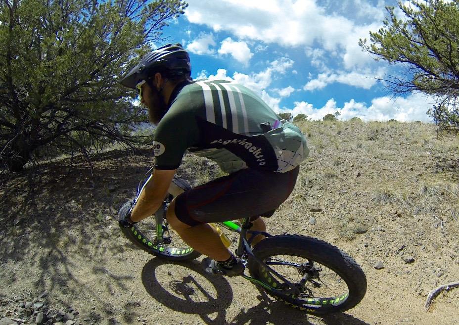 A cyclist navigating a dirt trail on a mountain bike, wearing a helmet and cycling gear. The background features a desert landscape with shrubs and a clear blue sky with scattered clouds. Arkansas Hills mountain bike trail.
