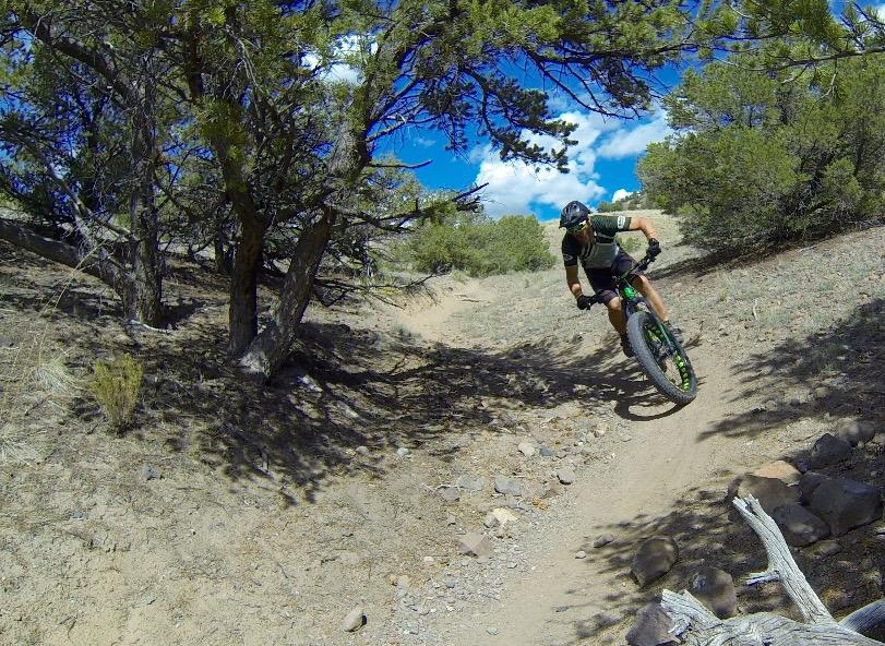 A cyclist riding a mountain bike on a dirt trail surrounded by trees. The rider is in a dynamic position, leaning into a turn, with a partly cloudy blue sky in the background. Arkansas Hills mountain bike trail.