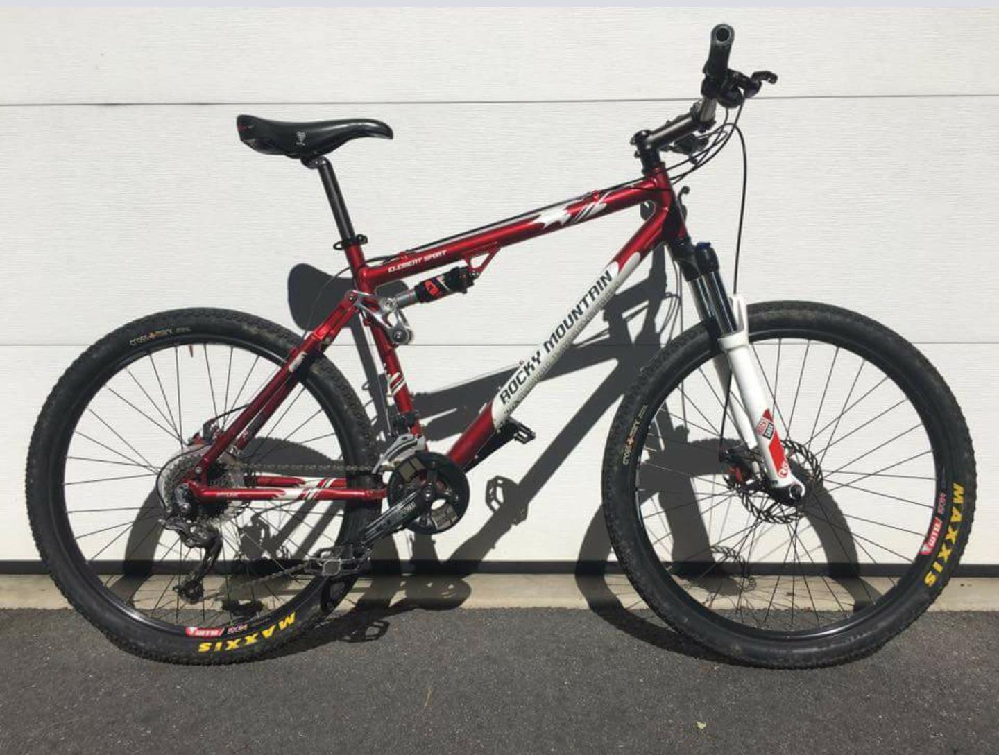 Rocky Mountain Element: A red and white mountain bike leaning against a light-colored wall, featuring thick tires with "Maxxis" branding, a dual suspension frame, and a black saddle. The bike is oriented to show its profile, highlighting its design and components.