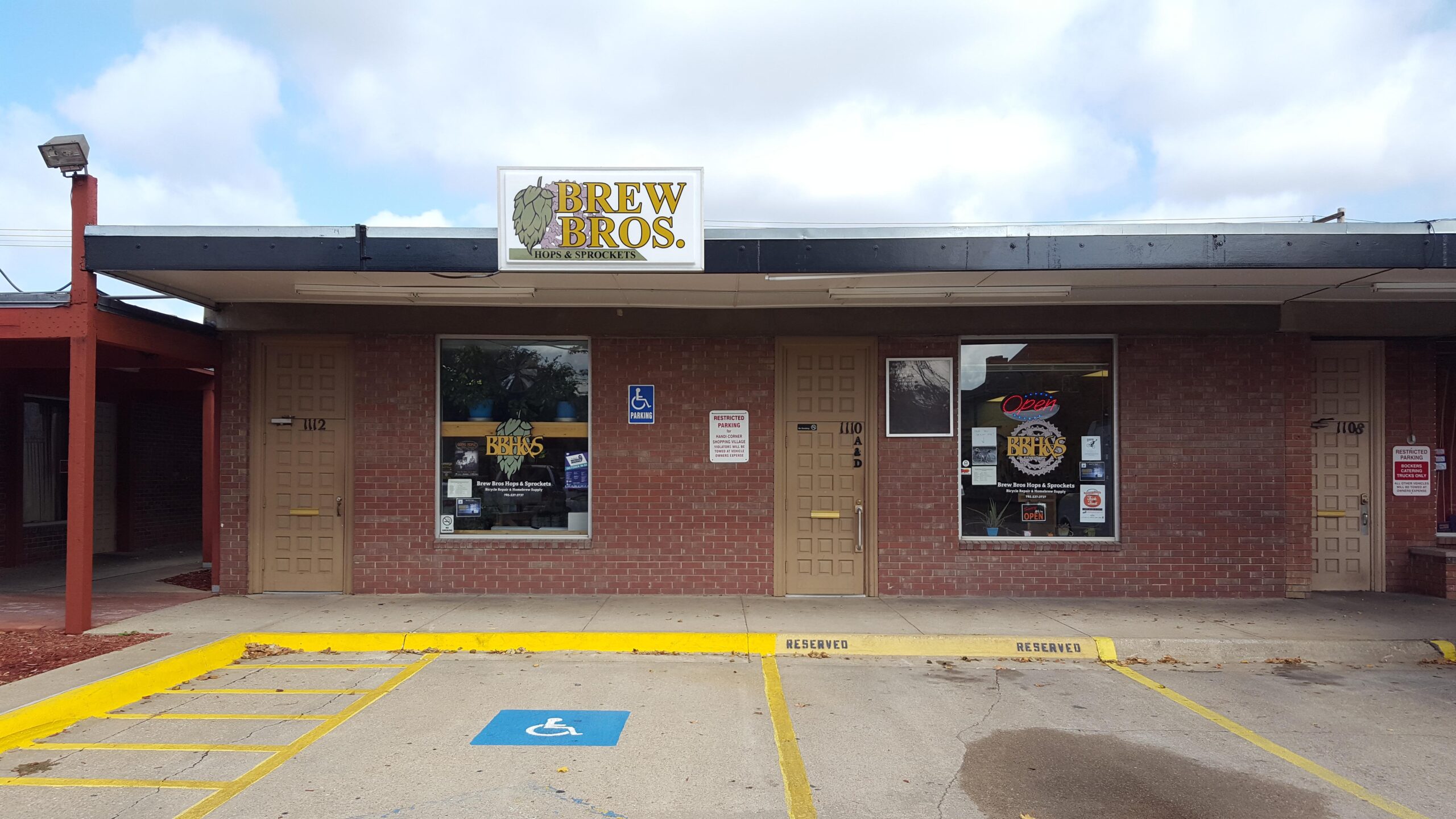 Exterior view of a storefront featuring the sign "Brew Bros. Hops & Sprockets." The building is made of red brick with a large window displaying the interior. There is a handicapped parking spot in front and a door with the number "110" next to it. The sky is partly cloudy.