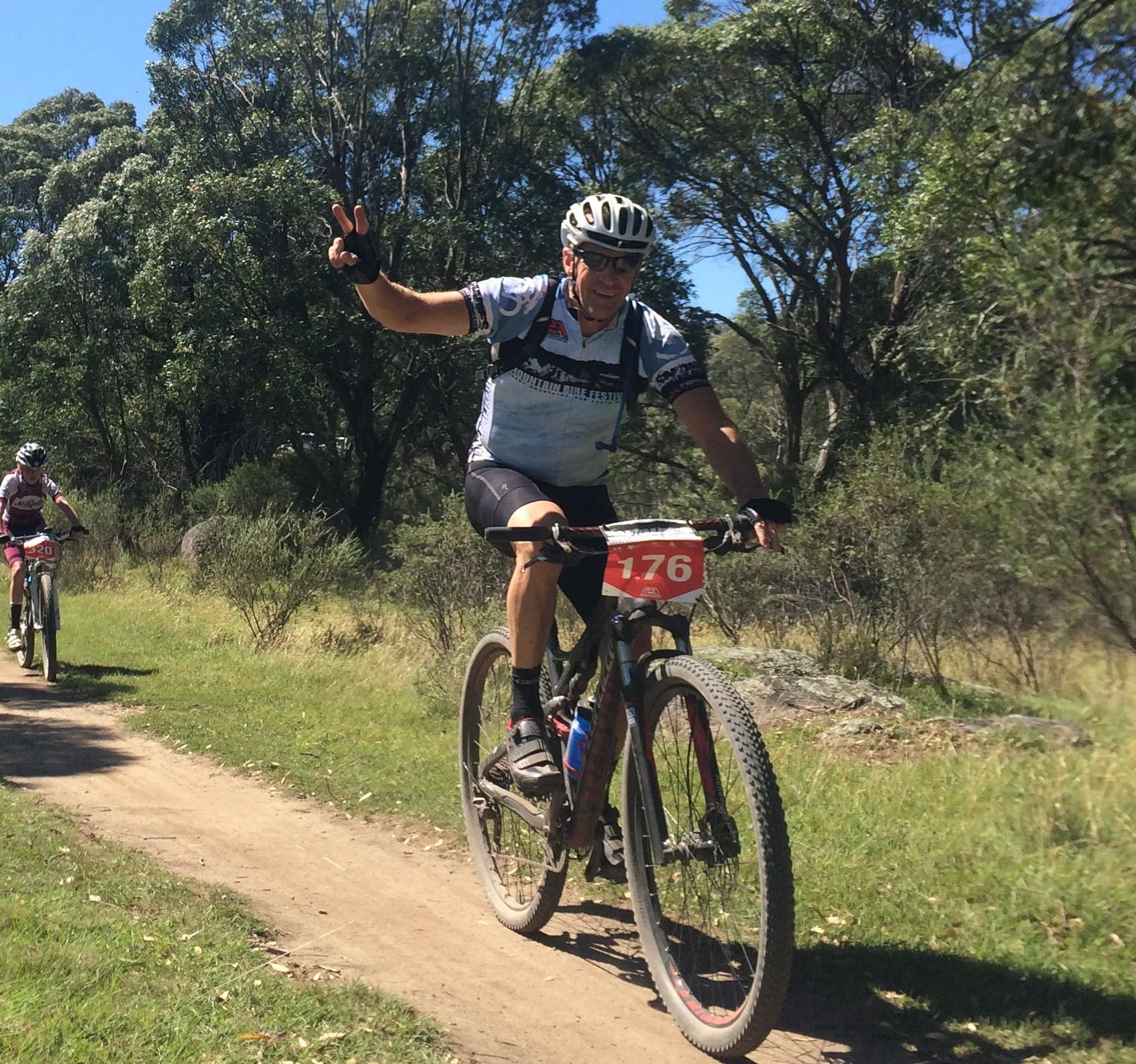 Specialized EPIC COMP CARBON: A cyclist wearing a blue and white jersey with the number 176 rides along a dirt path, smiling and giving a peace sign with one hand. Another cyclist in the background also rides on the trail, surrounded by green trees and shrubbery under a clear blue sky.