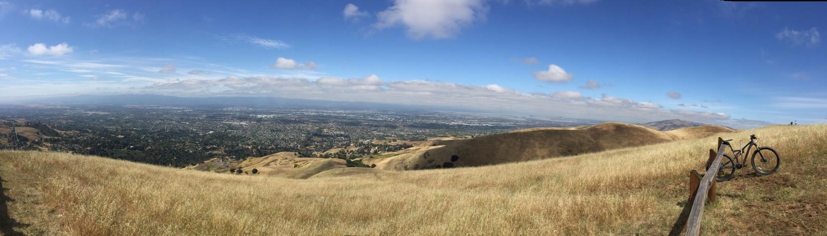 Specialized Camber Comp 29: A panoramic view of rolling hills covered in dry grass, with a scenic overlook of a valley and distant mountains under a partly cloudy sky. In the foreground, a black bicycle stands next to a wooden fence, with the expansive landscape stretching out behind it.