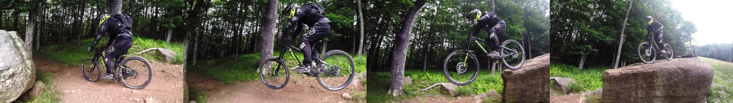 A mountain biker in protective gear jumps off a rocky ledge on a forested trail, showcasing action and skill while navigating the challenging terrain. The sequence captures the bike in mid-air, highlighting the rider's athleticism against a backdrop of lush greenery and trees. Snowshoe Bike Park mountain bike trail.