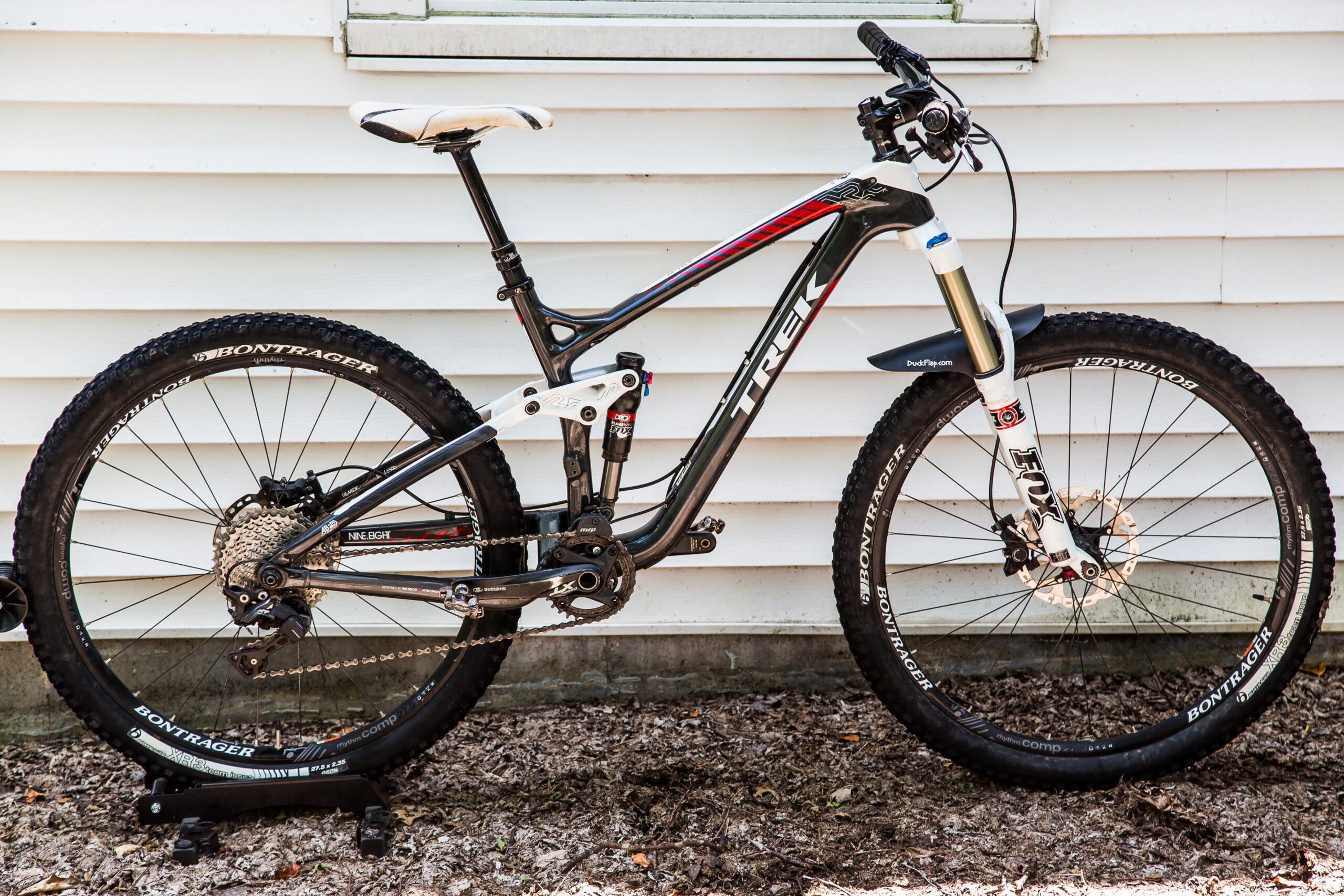 Trek Remedy 9.8 27.5/650b: Mountain bike leaning against a white wooden wall, featuring a sleek, black and white frame with red accents. The bike is equipped with Bontrager tires, a front suspension fork, and a visible drivetrain with gears and a rear derailleur. The ground is covered in leaves and dirt, indicating an outdoor setting.