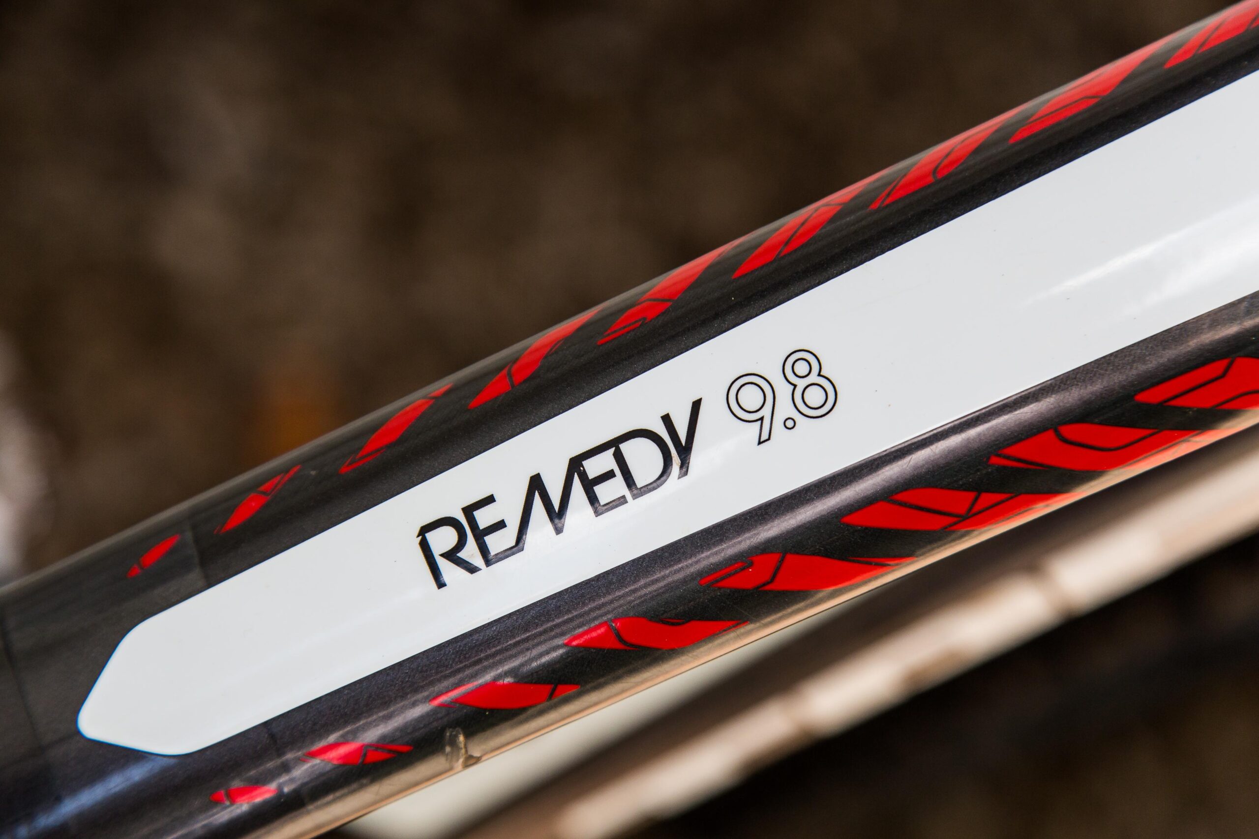 Trek Remedy 9.8 27.5/650b: Close-up view of a mountain bike frame featuring the model name "REMEDY 9.8" prominently displayed in bold black letters on a white background, surrounded by a graphic design with red accents on a black surface. The image captures the sleek detailing of the bike's frame.