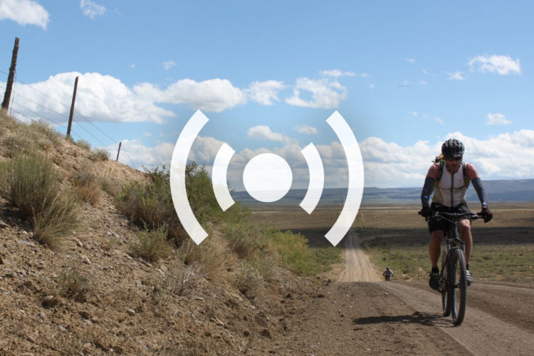 A cyclist riding on a dirt trail through a rural landscape with sparse vegetation, set against a blue sky with fluffy white clouds. In the background, another cyclist can be seen in the distance. A graphic symbol representing sound or broadcasting is overlayed in the center of the image.