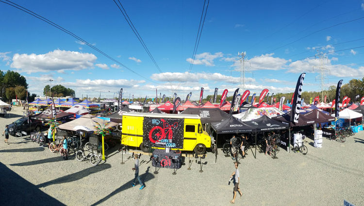 A panoramic view of a vibrant outdoor event featuring numerous vendor tents and flags, showcasing various cycling brands. The scene includes colorful tents, displayed bicycles, and attendees walking around, all under a clear blue sky with scattered clouds.