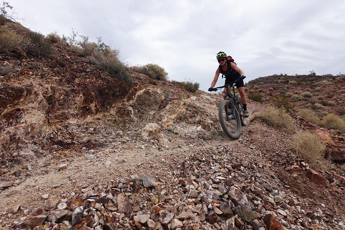 A mountain biker riding on a rocky trail, navigating through the rough terrain, with a backdrop of hills and cloudy skies. The biker is wearing a helmet and is equipped with a backpack, showcasing an adventurous outdoor scene. Bootleg Canyon mountain bike trail.