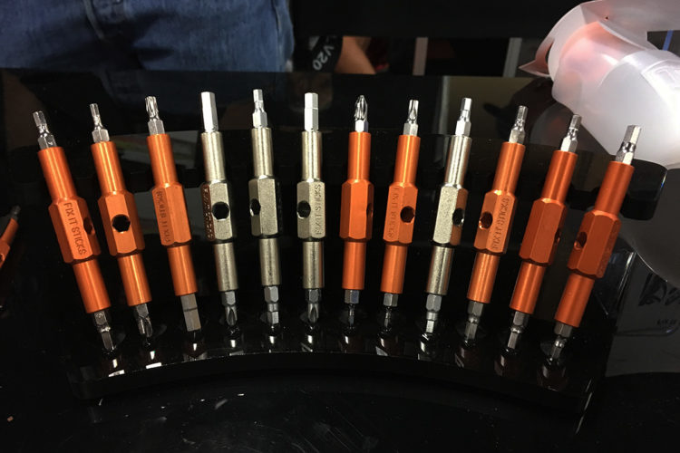 A display of a variety of multi-tool screwdriver bits arranged in a curved holder. The bits feature different heads, including flat and Phillips types, with some in orange and silver metallic finishes. The holder is black and glossy, reflecting light. In the background, part of a person's jeans is visible.