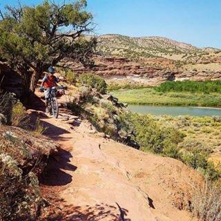 A mountain biker riding along a rocky path with a scenic view of a river and green vegetation in the background, under a clear blue sky. Kokopelli Area Trails mountain bike trail.
