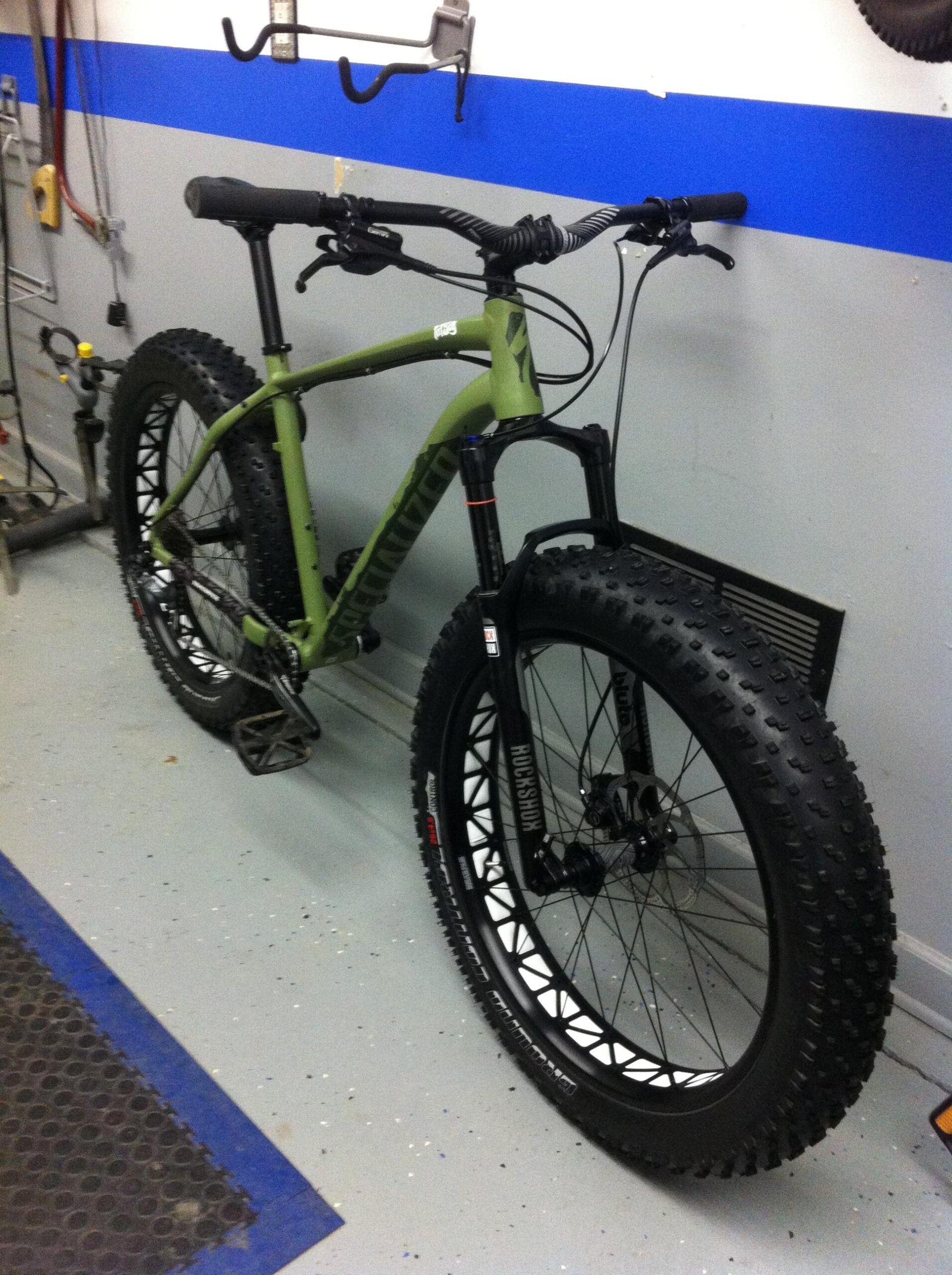 Specialized Stumpjumper FSR comp carbon 650B: A green fat bike with wide tires and a sturdy frame is positioned in a garage, with a blue stripe on the wall behind it. The bike features thick, knobby tires ideal for off-road riding and has a front suspension fork. Various tools and equipment are visible in the background, indicating a workspace for bike maintenance.