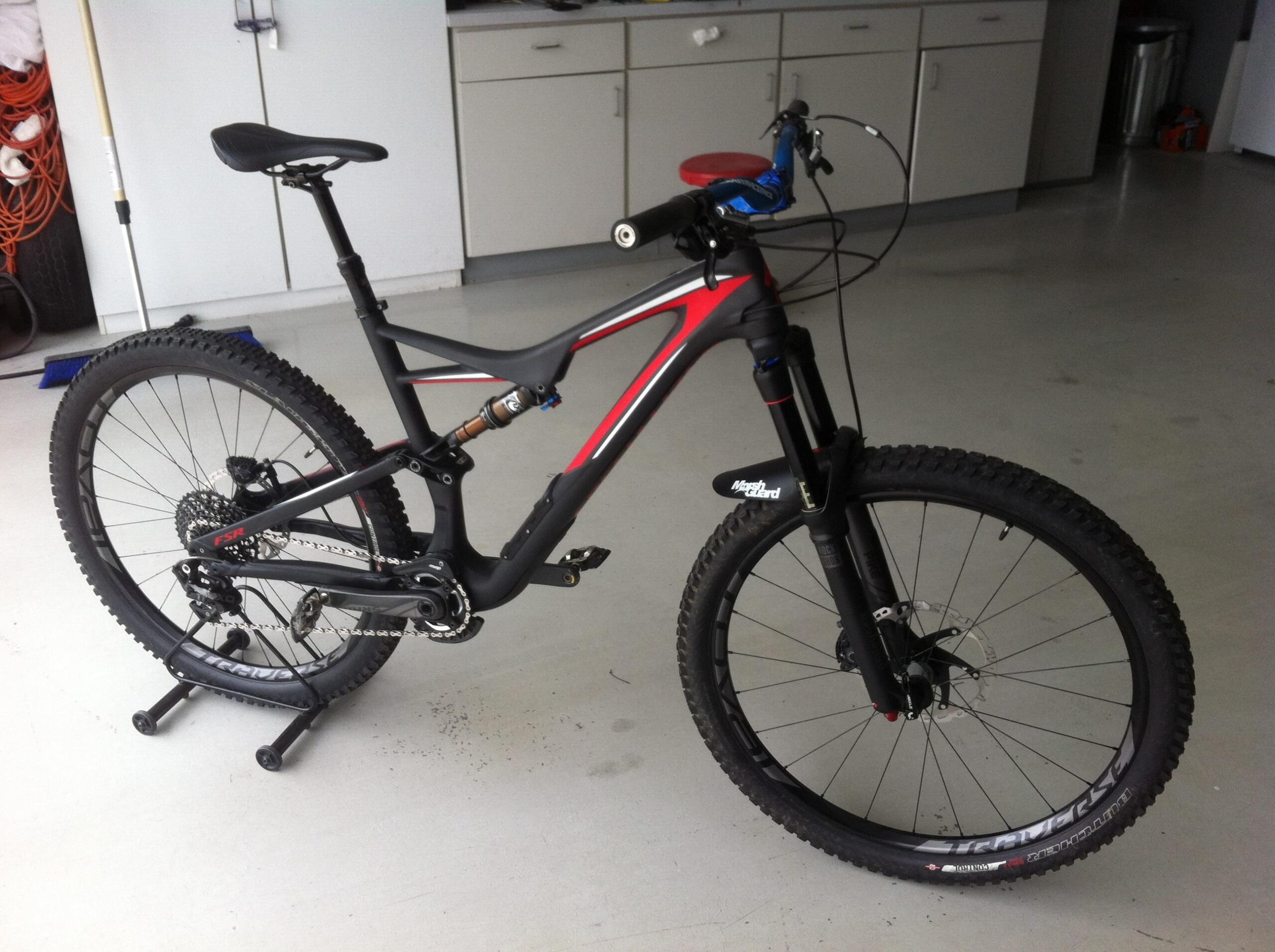 Specialized Stumpjumper FSR comp carbon 650B: A black and red mountain bike displayed in a garage, positioned on a bike stand. The bike features thick tires, a suspension fork, and gear components visible on the frame. The background includes cabinets and cleaning tools.