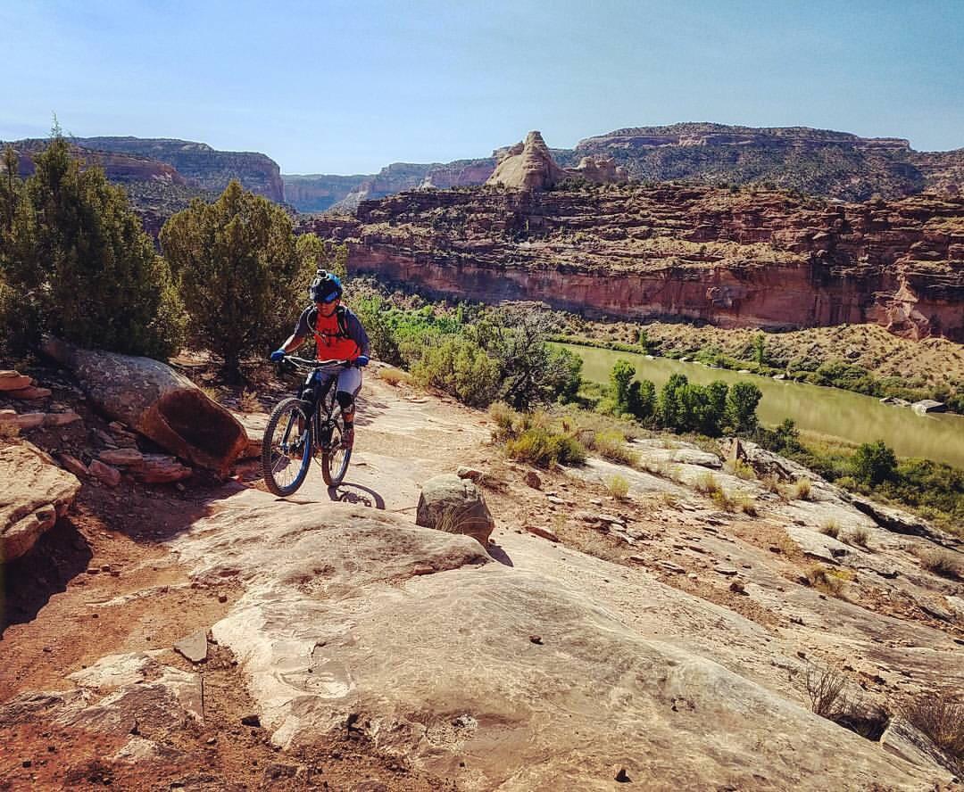 A mountain biker navigates a rocky trail in a scenic landscape, featuring cliffs and greenery, with a river visible in the background under a clear blue sky. Mary's Loop / Horsethief Bench mountain bike trail.