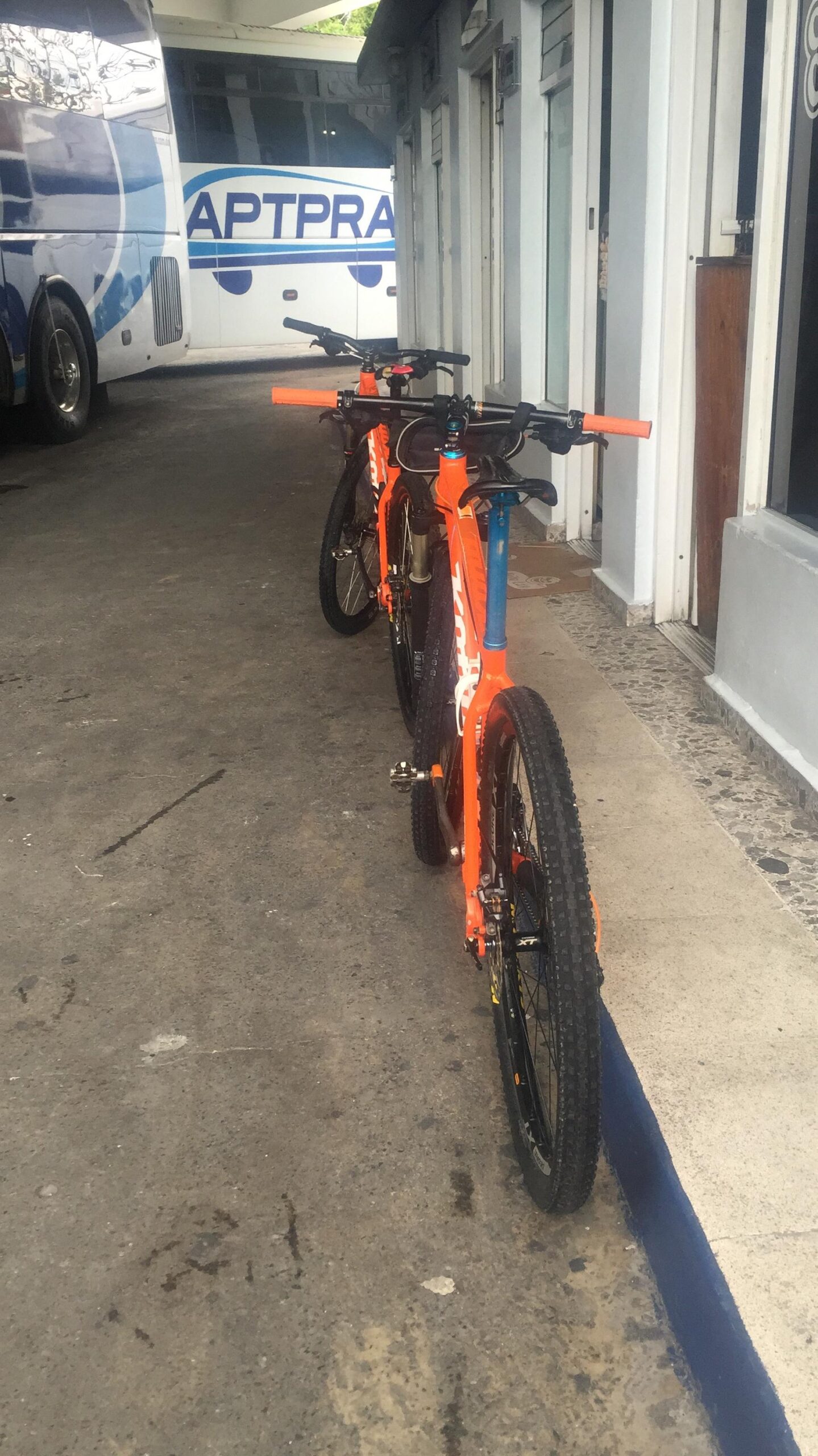 Kona Kahuna: A pair of orange mountain bikes parked along a narrow walkway next to a bus. The bikes are positioned on the left side of the image, with the bus visible in the background. The scene features a concrete surface and buildings to the right, creating a casual urban setting.