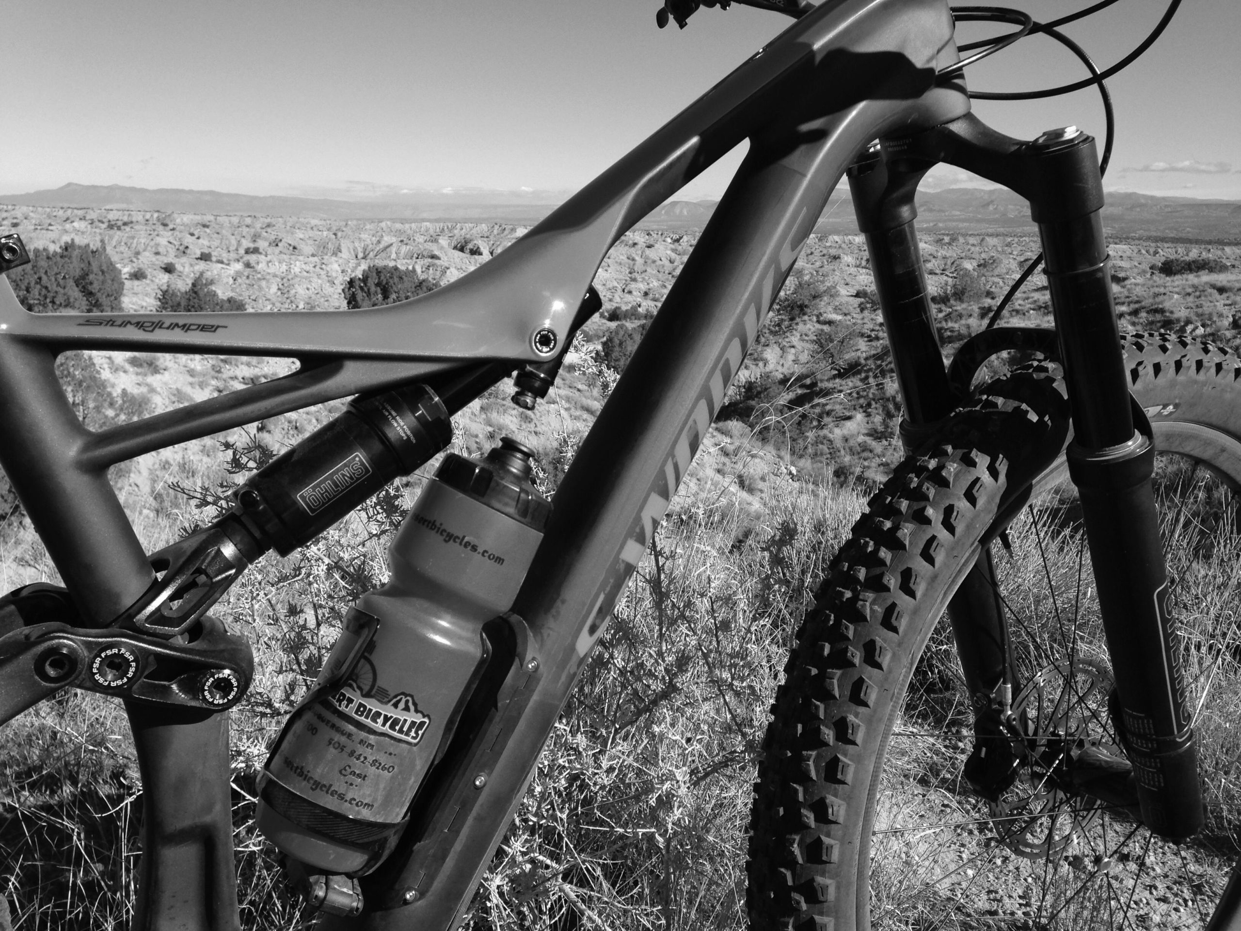 Close-up view of a mountain bike frame, featuring the brand name "Stumpjumper," with a shock absorber and a water bottle attached to the frame. The background reveals a vast, rugged landscape with hills and distant mountains. The image is in black and white. Mariposa Fat Bike Trails mountain bike trail.
