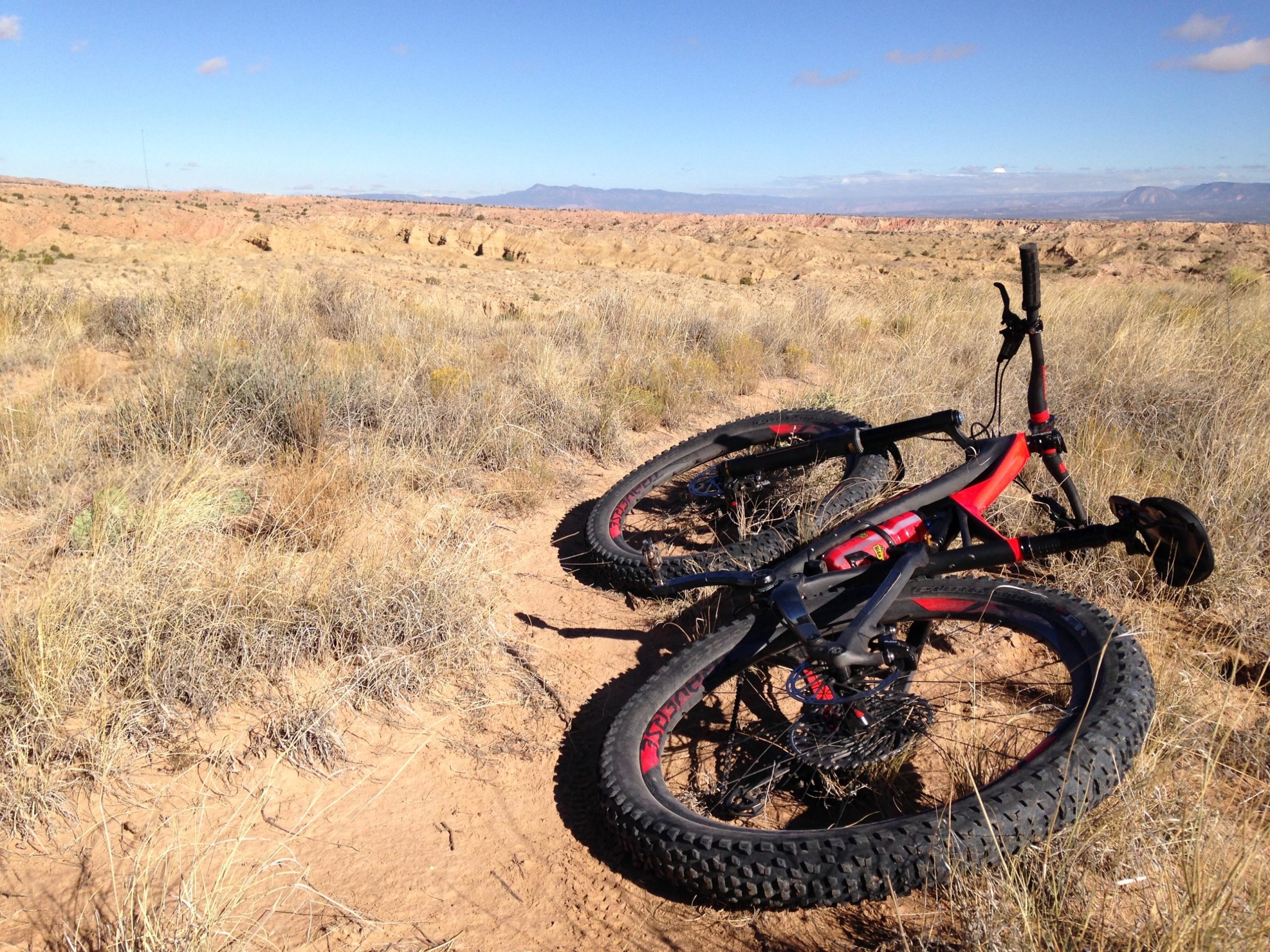 A pair of mountain bikes resting on a sandy and grassy terrain, with a scenic backdrop of a rugged landscape and blue sky. The bikes have thick tires designed for off-road cycling, and their red and black frames suggest they are ready for adventure in a natural outdoor setting. Mariposa Fat Bike Trails mountain bike trail.