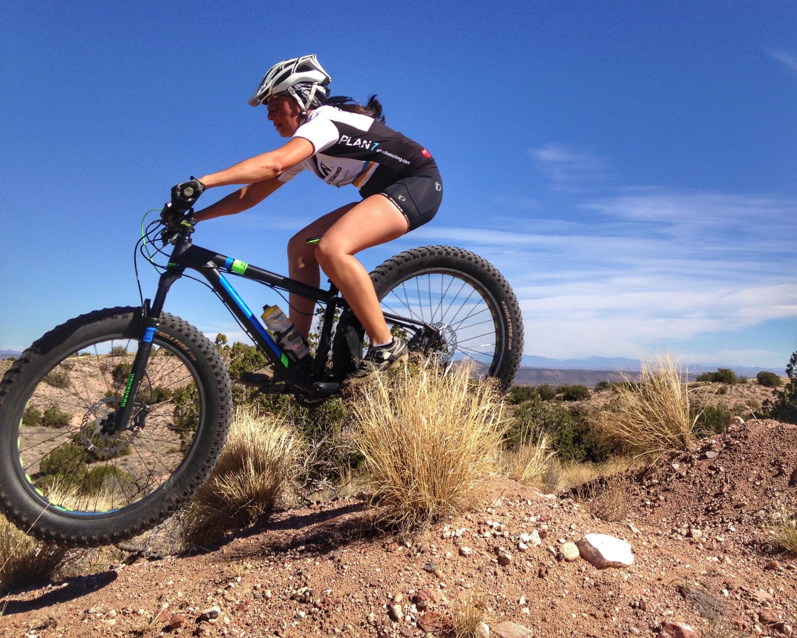 A woman in cycling gear rides a mountain bike over a rocky terrain, captured mid-air as she jumps off a ledge. The background features a clear blue sky and sparse vegetation, highlighting an outdoor mountain biking environment. Mariposa Fat Bike Trails mountain bike trail.