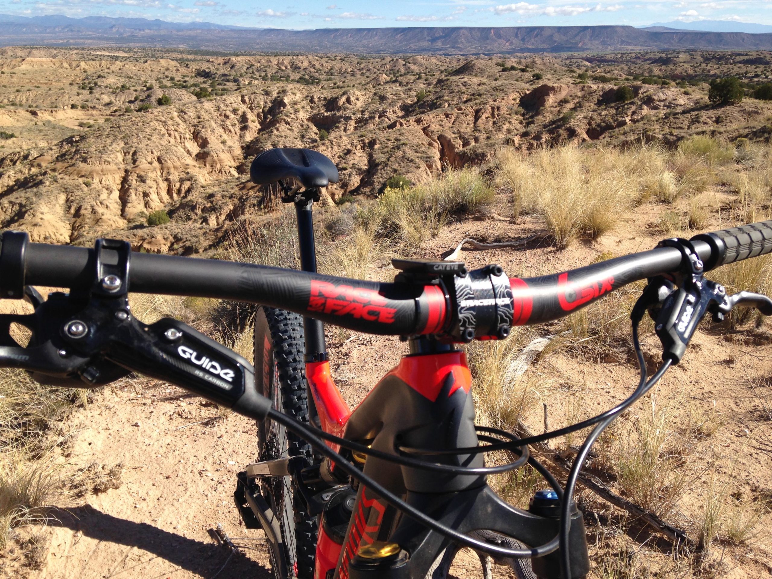 Close-up view of a mountain bike handlebars positioned on a rocky terrain with a vast landscape in the background, featuring rolling hills and distant mountains under a blue sky. Mariposa Fat Bike Trails mountain bike trail.