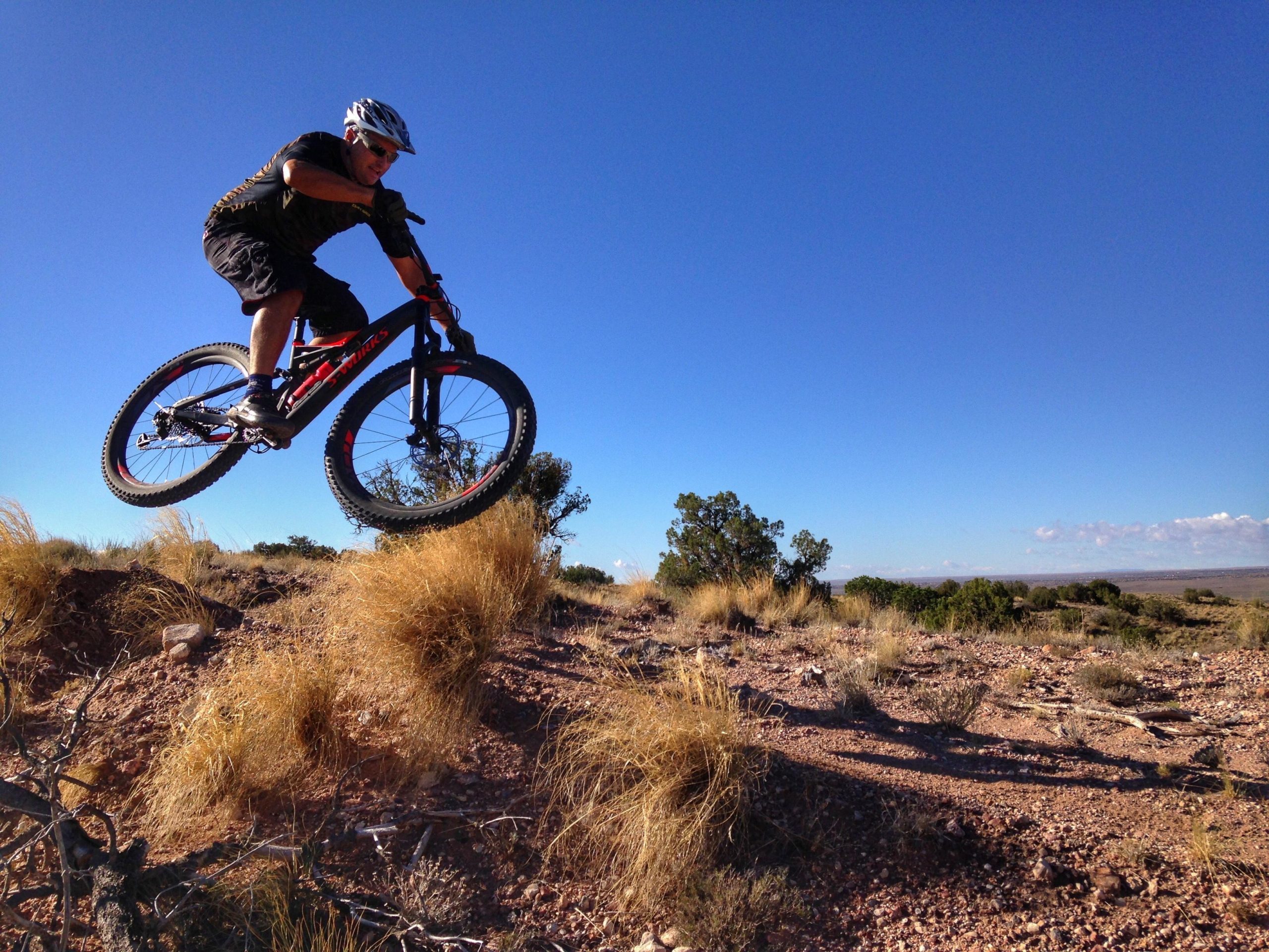 A mountain biker performing a jump over a rocky terrain, set against a clear blue sky, with dry grasses and sparse vegetation in the foreground. Mariposa Fat Bike Trails mountain bike trail.