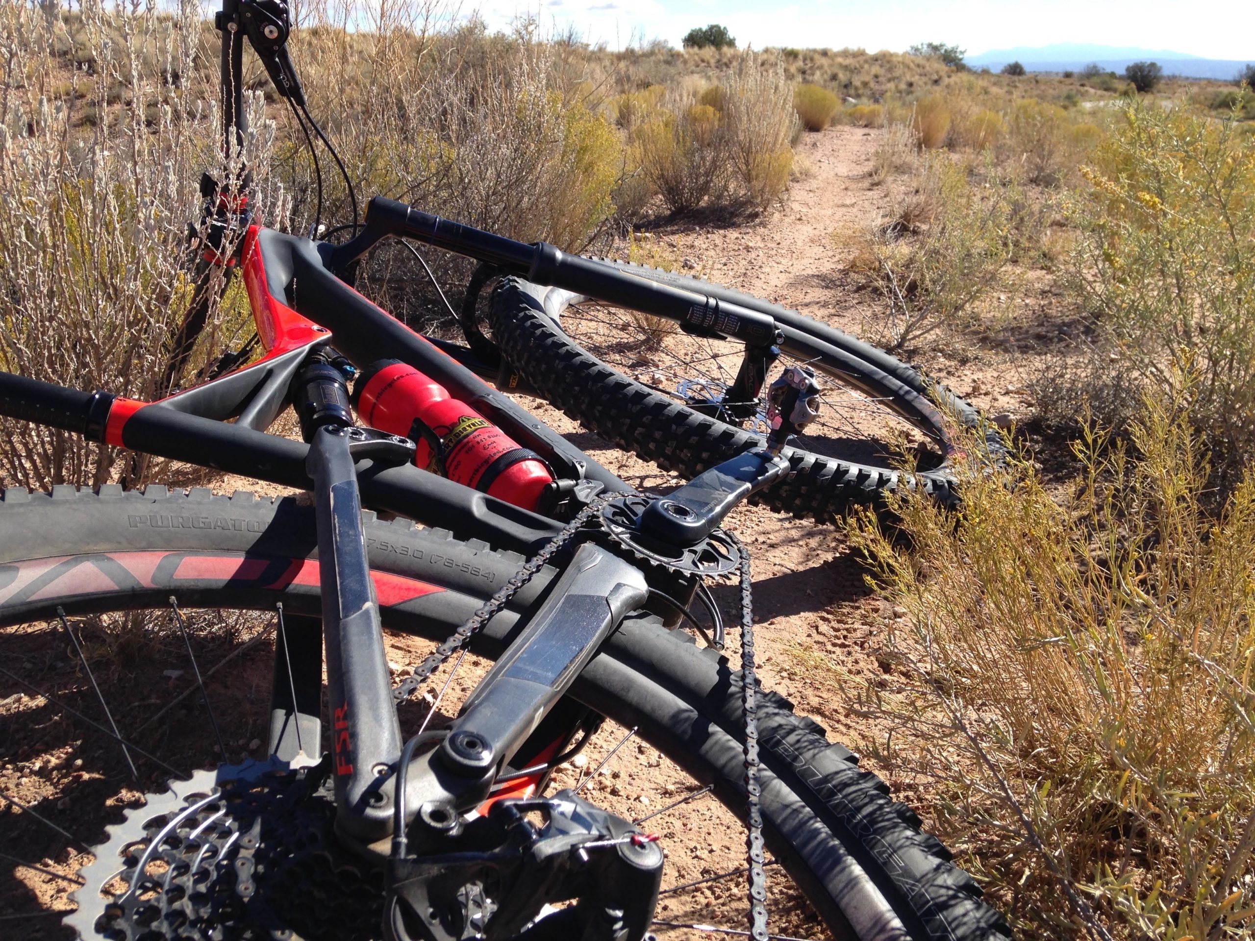 Mountain bike resting on a gravel path surrounded by desert vegetation. The bike features a black frame with red accents, and its wheels are partially in the frame, showing off rugged tires. The background includes sparse shrubs and distant hills under a clear sky. Parkway Fatbike trail mountain bike trail.