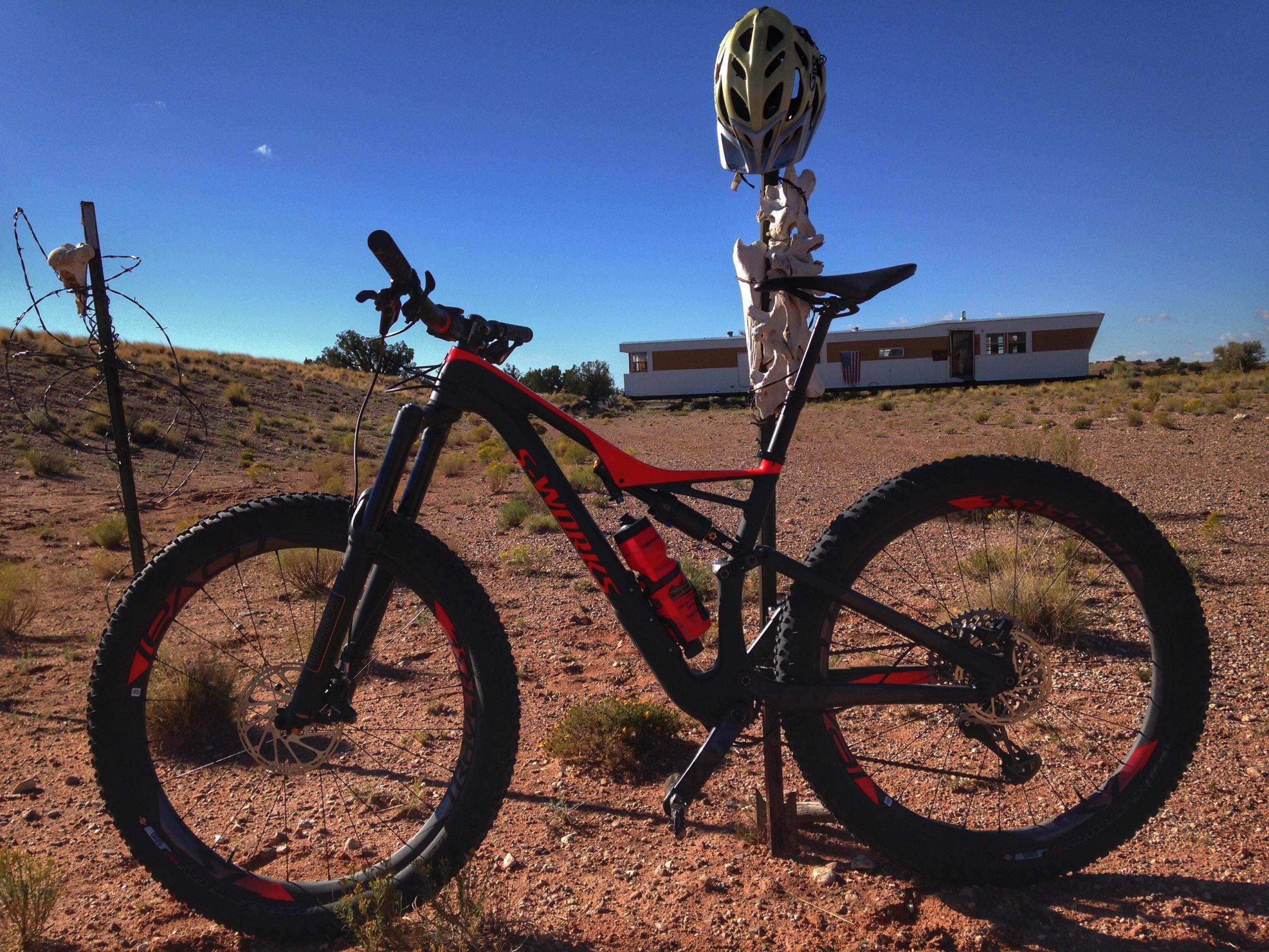 A mountain bike parked on a dirt trail with a blue sky in the background. In the distance, a mobile home is visible, and there is a barbed wire fence with a skull decoration nearby. The scene captures a remote and rustic outdoor setting. Mariposa Fat Bike Trails mountain bike trail.