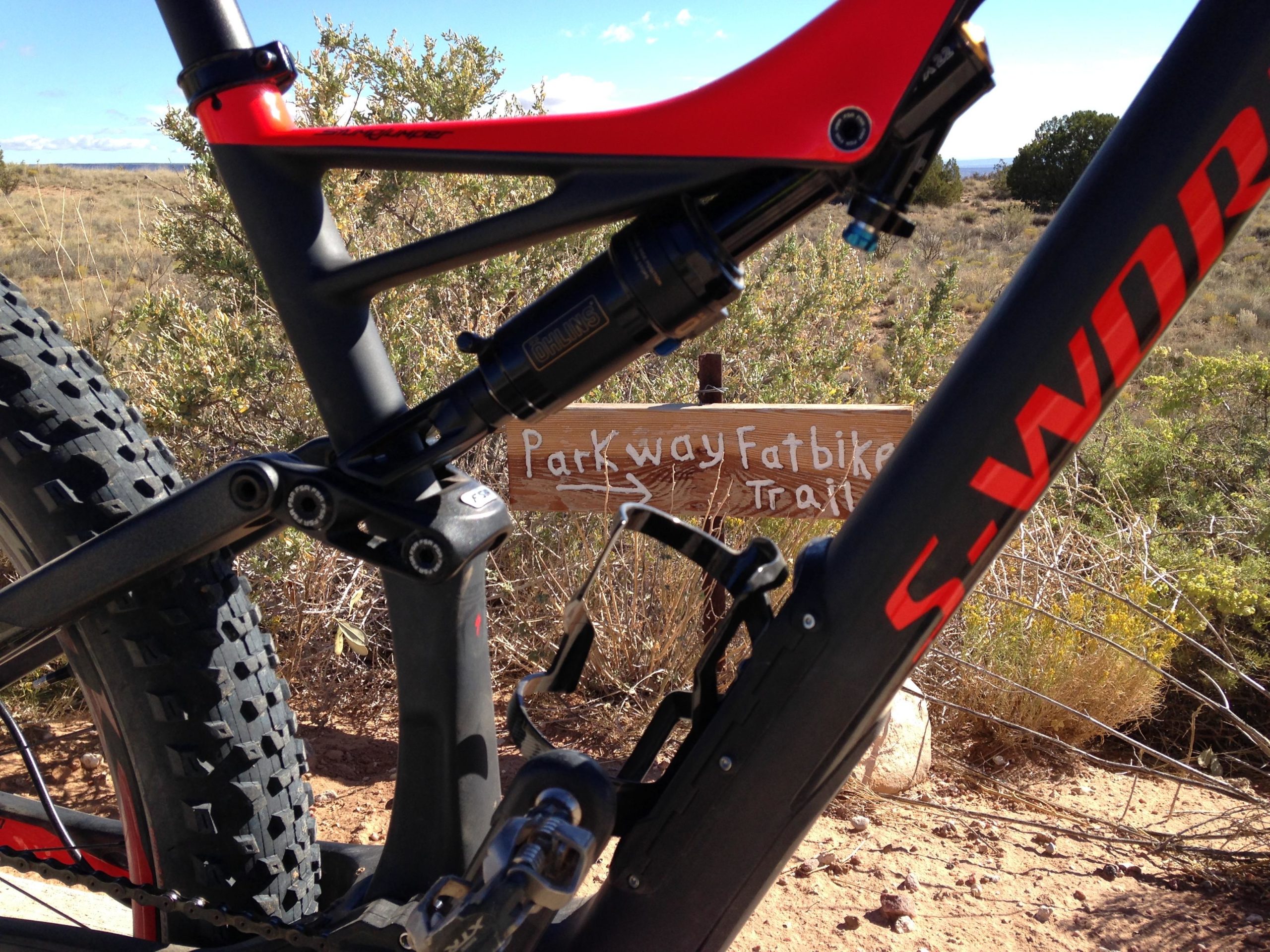 Close-up view of a fatbike with a vibrant red and black frame, partially obscured by shrubbery, alongside a wooden sign that reads "Parkway Fatbike Trail." The background showcases an open terrain under a clear blue sky. Parkway Fatbike trail mountain bike trail.