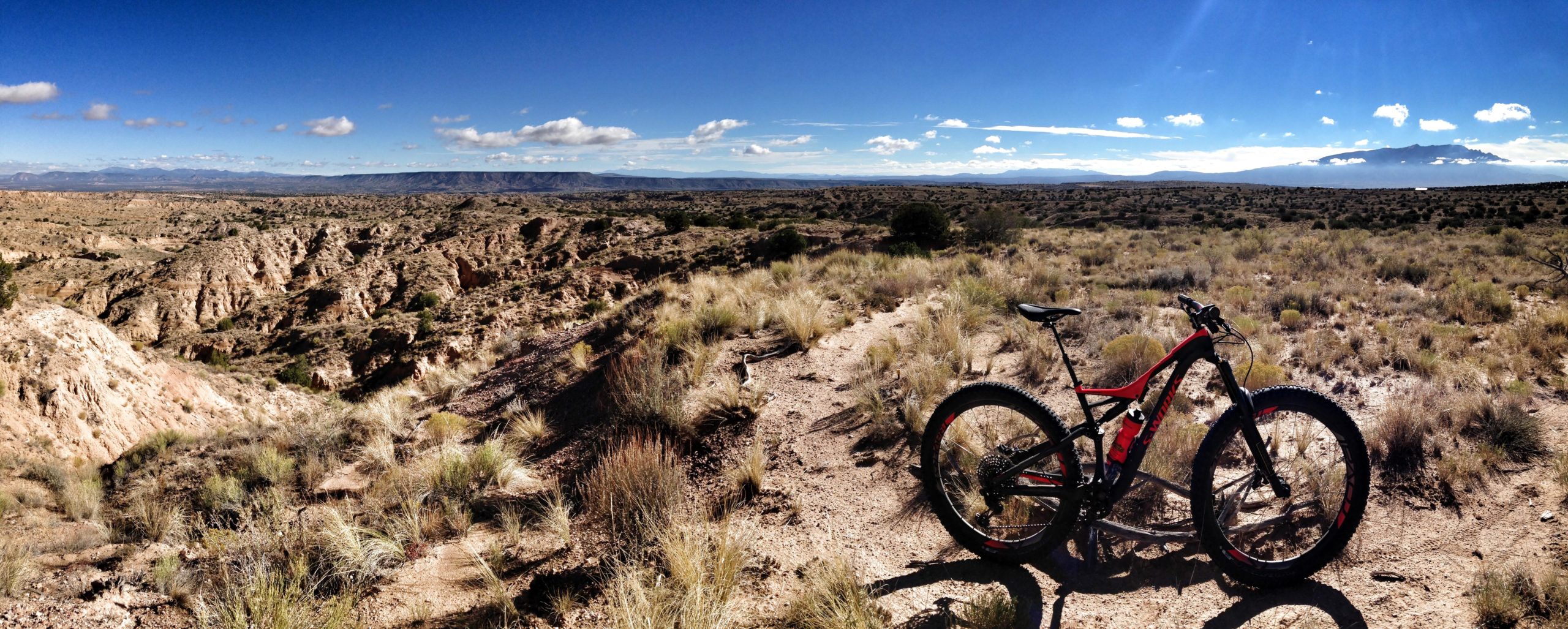 A mountain bike resting on a rocky trail, overlooking a vast desert landscape with rolling hills and distant mountains under a clear blue sky with a few fluffy clouds. The foreground features dry grasses and rugged terrain typical of a desert environment. Mariposa Fat Bike Trails mountain bike trail.