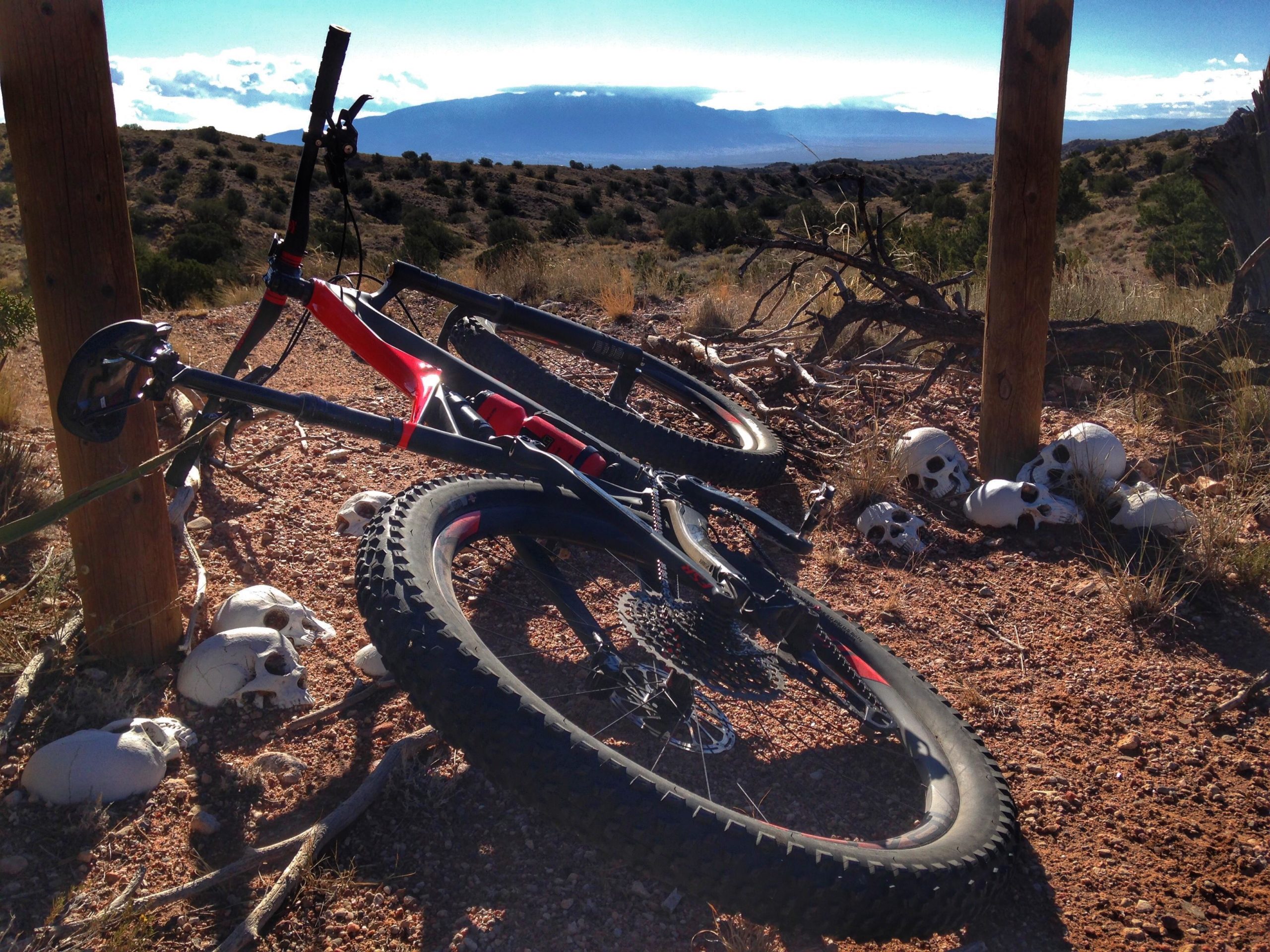 A mountain bike resting against a wooden post in a rocky outdoor landscape, with several white skulls scattered on the ground nearby. The scene is illuminated by natural sunlight, and the background features rolling hills and a cloudy sky. Mariposa Fat Bike Trails mountain bike trail.