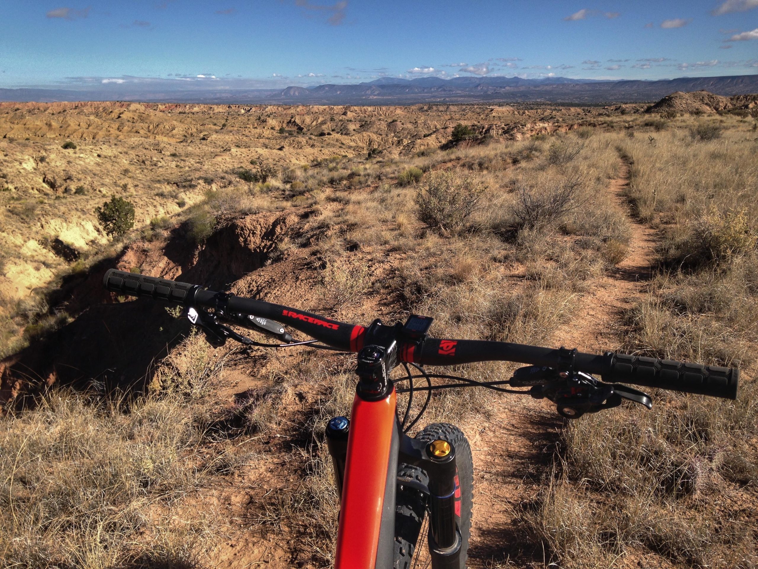 Mountain bike handlebars in the foreground, overlooking a scenic, rugged landscape with rocky terrain and low vegetation under a clear blue sky. A dirt trail winds through the scenery, leading into the distance with mountains on the horizon. Mariposa Fat Bike Trails mountain bike trail.