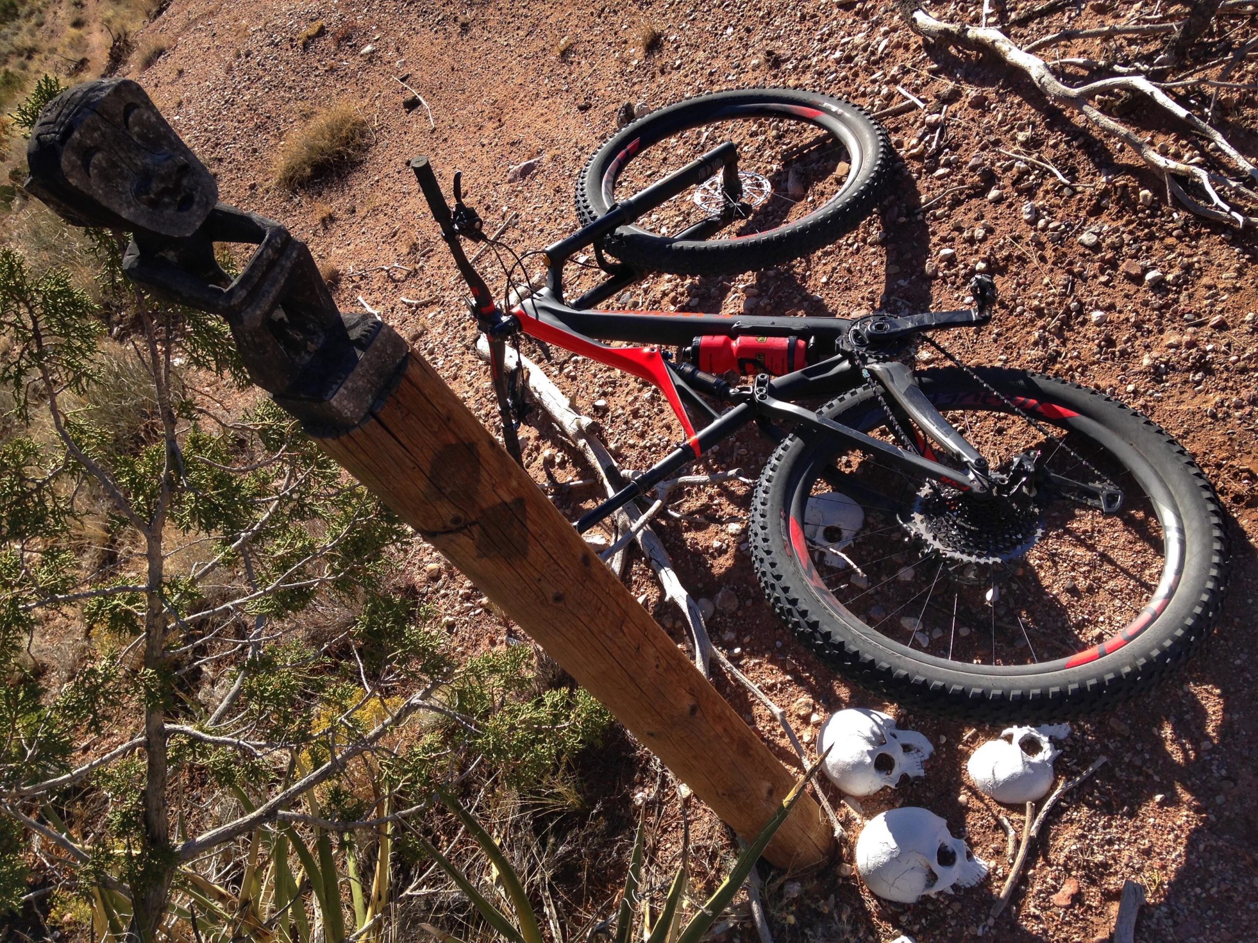 A mountain bike rests on the ground in a rugged, desert landscape, beside a wooden post topped with a carved figure's head. Nearby, three white skulls are scattered on the dirt, surrounded by sparse vegetation and scattered branches. Mariposa Fat Bike Trails mountain bike trail.