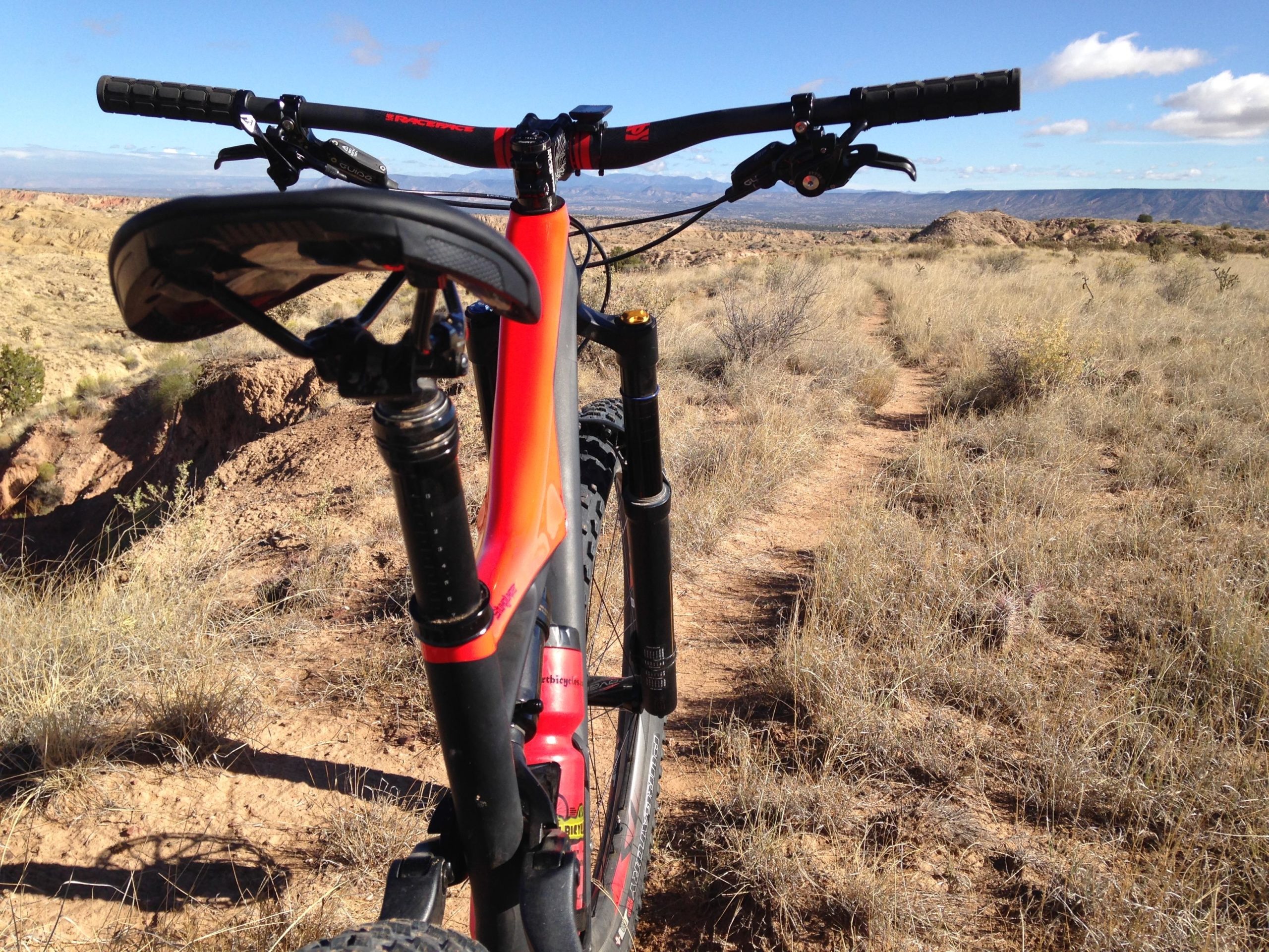 A mountain bike is positioned on a dirt trail surrounded by dry grass and rugged terrain, with a clear blue sky above and distant mountains in the background. The focus is on the bike's saddle and handlebars, highlighting its design and structure. Mariposa Fat Bike Trails mountain bike trail.