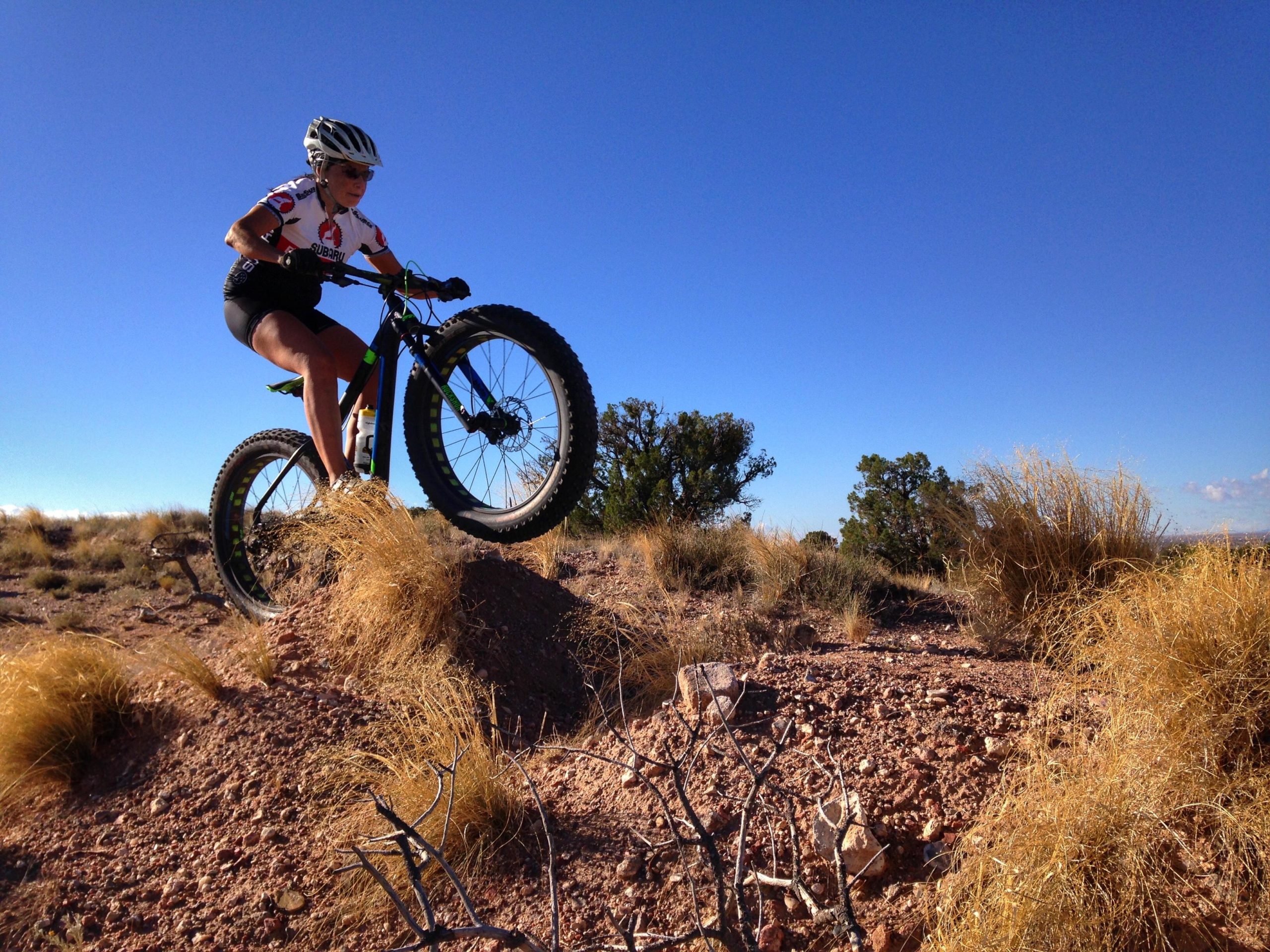 A person riding a fat tire mountain bike performs a jump over a small mound of earth in a desert landscape. The scene features sparse vegetation and blue skies in the background. The cyclist is wearing a helmet and a sports jersey, showcasing an active outdoor adventure. Mariposa Fat Bike Trails mountain bike trail.