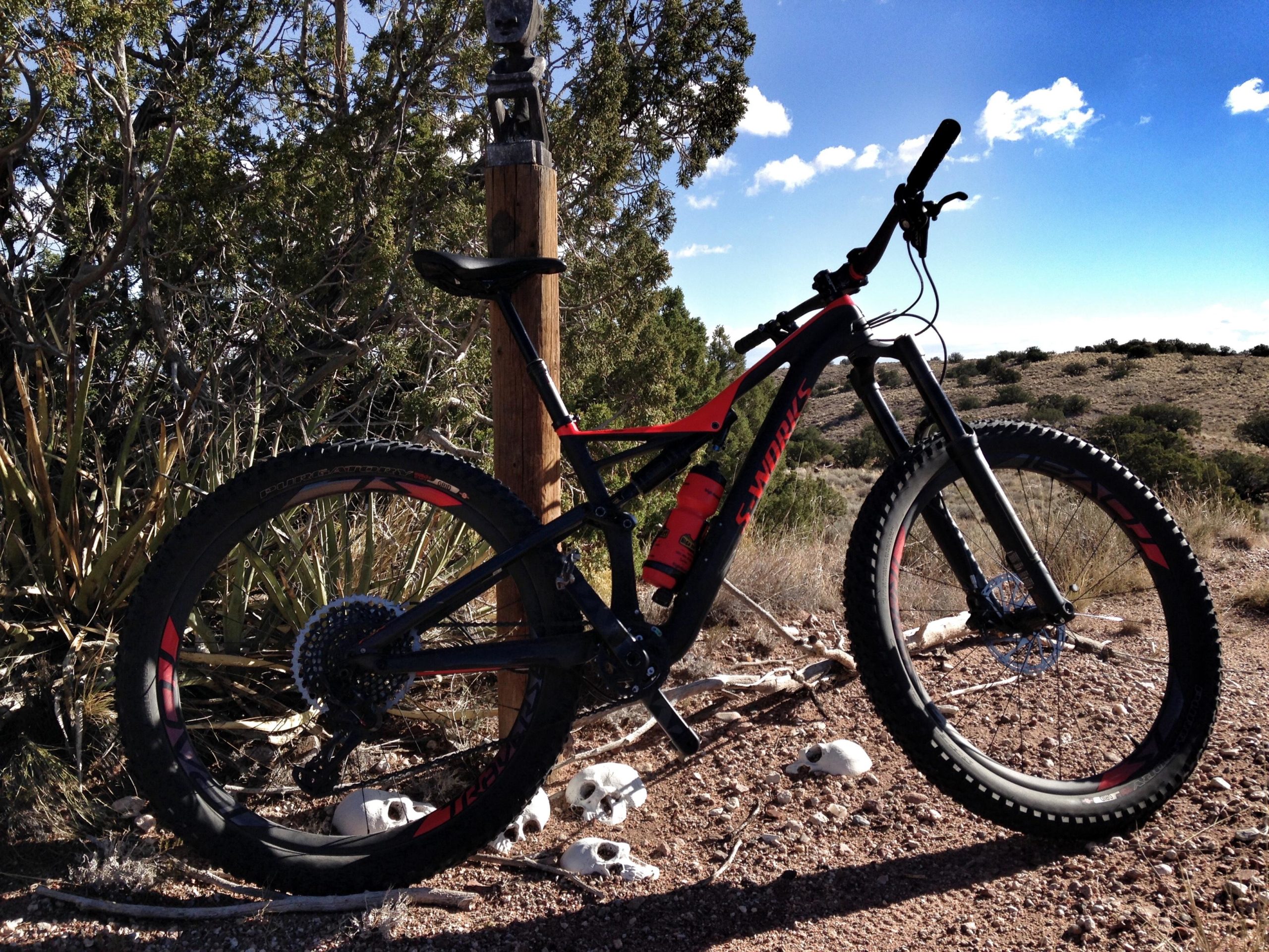 A mountain bike leaning against a wooden post in a desert landscape, with scrub and rocks in the background. The bike features thick, knobby tires and a bright red and black color scheme, and there are small skulls on the ground nearby. The sky is clear with a few scattered clouds. Mariposa Fat Bike Trails mountain bike trail.
